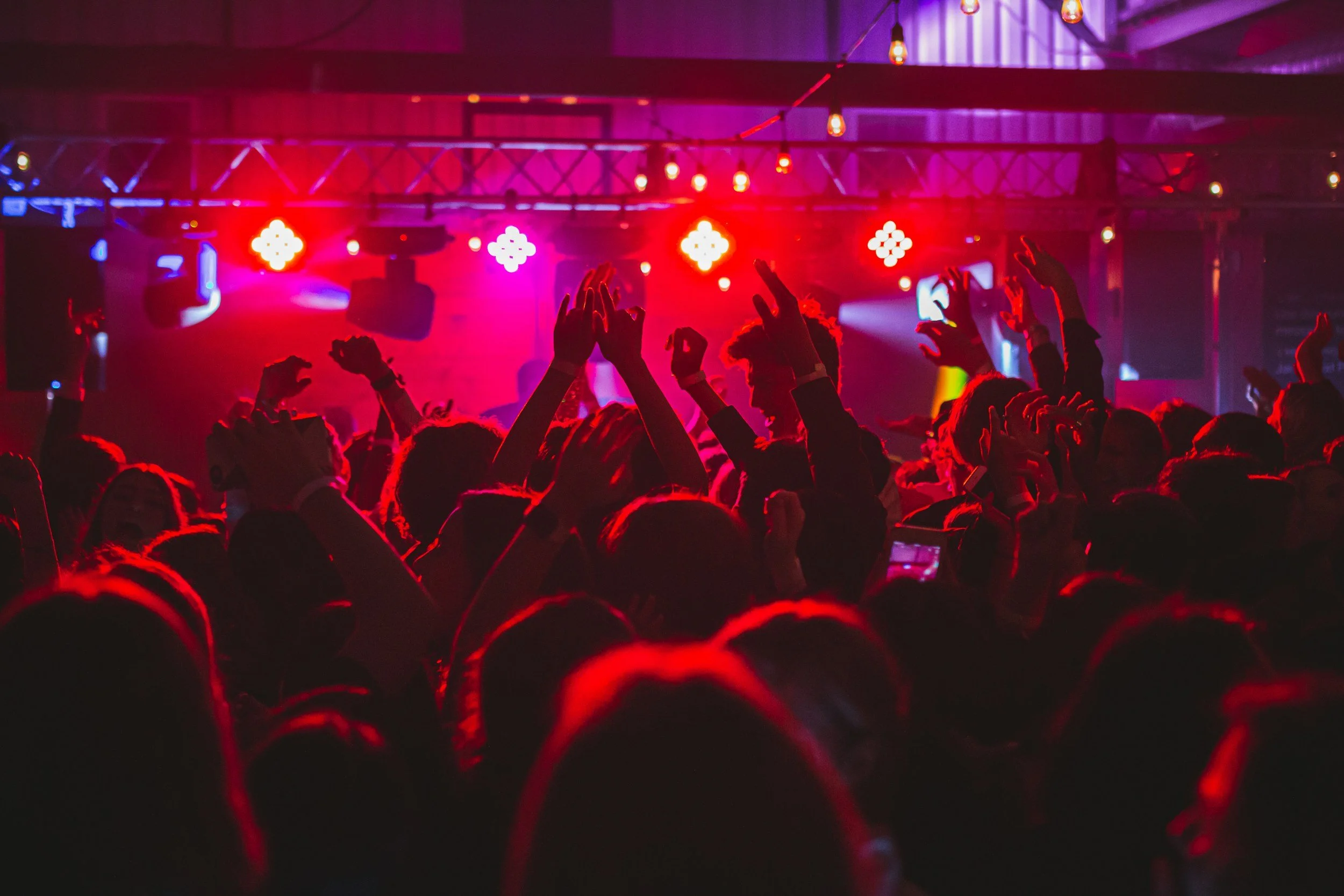 Crowd of people attending a concert or dance party with colorful stage lights and raised hands.