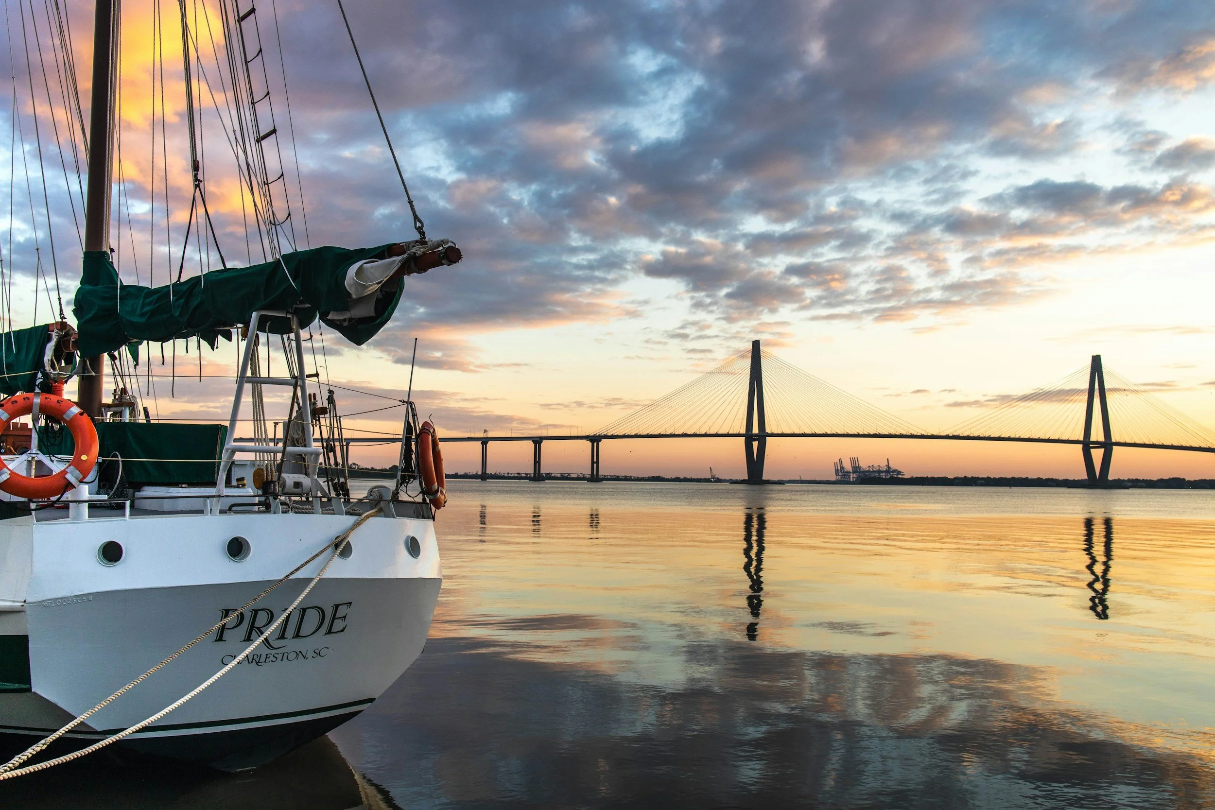 A sailboat named 'PRIDE' docked on calm water during sunset, with a bridge and industrial cranes in the background under a partly cloudy sky with soft orange and blue hues.
