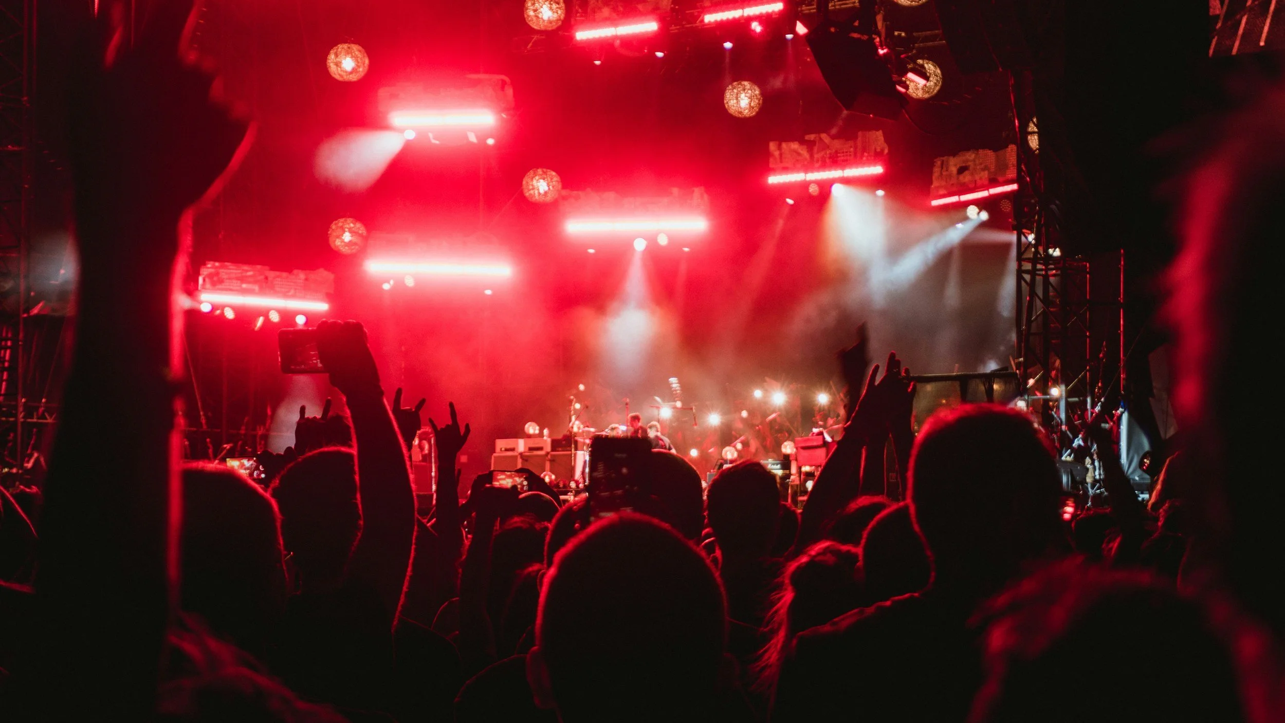 A concert scene with a crowd at night, illuminated by red stage lights, with hands in the air, capturing a live performance.