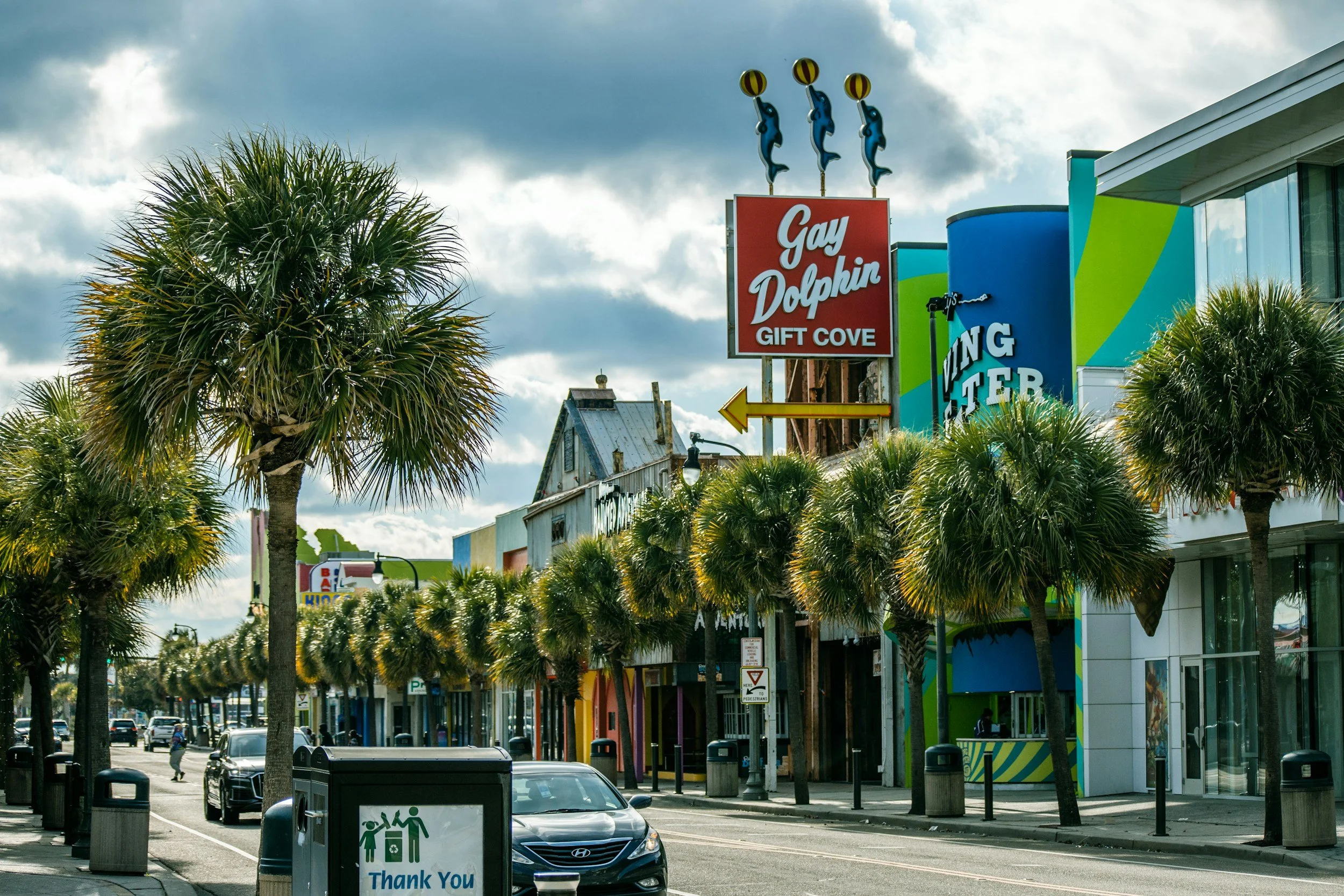 Street view at Gift Cove with palm trees, colorful buildings, and a sign reading 'Gay Dolphin Gift Cove' topped with three dolphins holding balls.