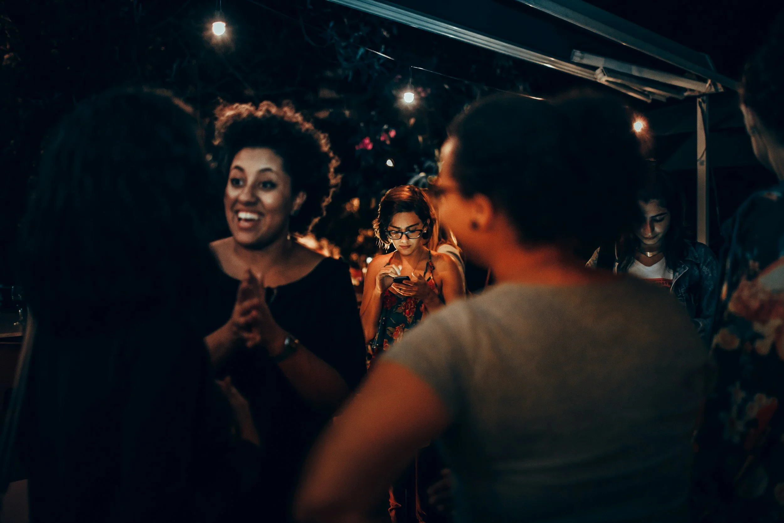 People gathered outdoors at night with string lights overhead, some are talking, and a woman in the center is smiling and laughing.