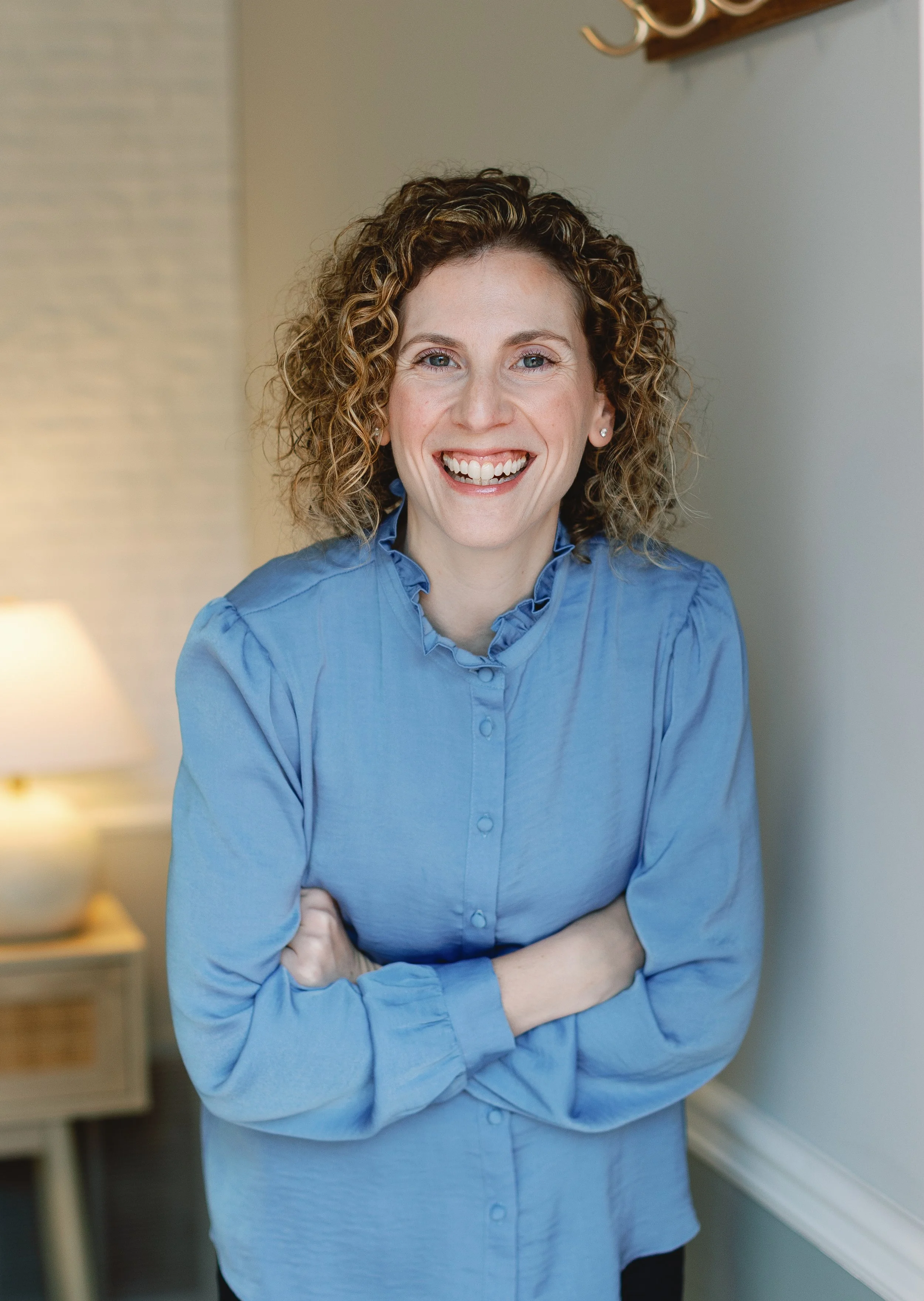 A woman with curly hair wearing a light blue blouse, smiling with arms crossed, standing in a room with neutral-colored walls. Psychiatric Care - Amy Zoller Lee - PA-C