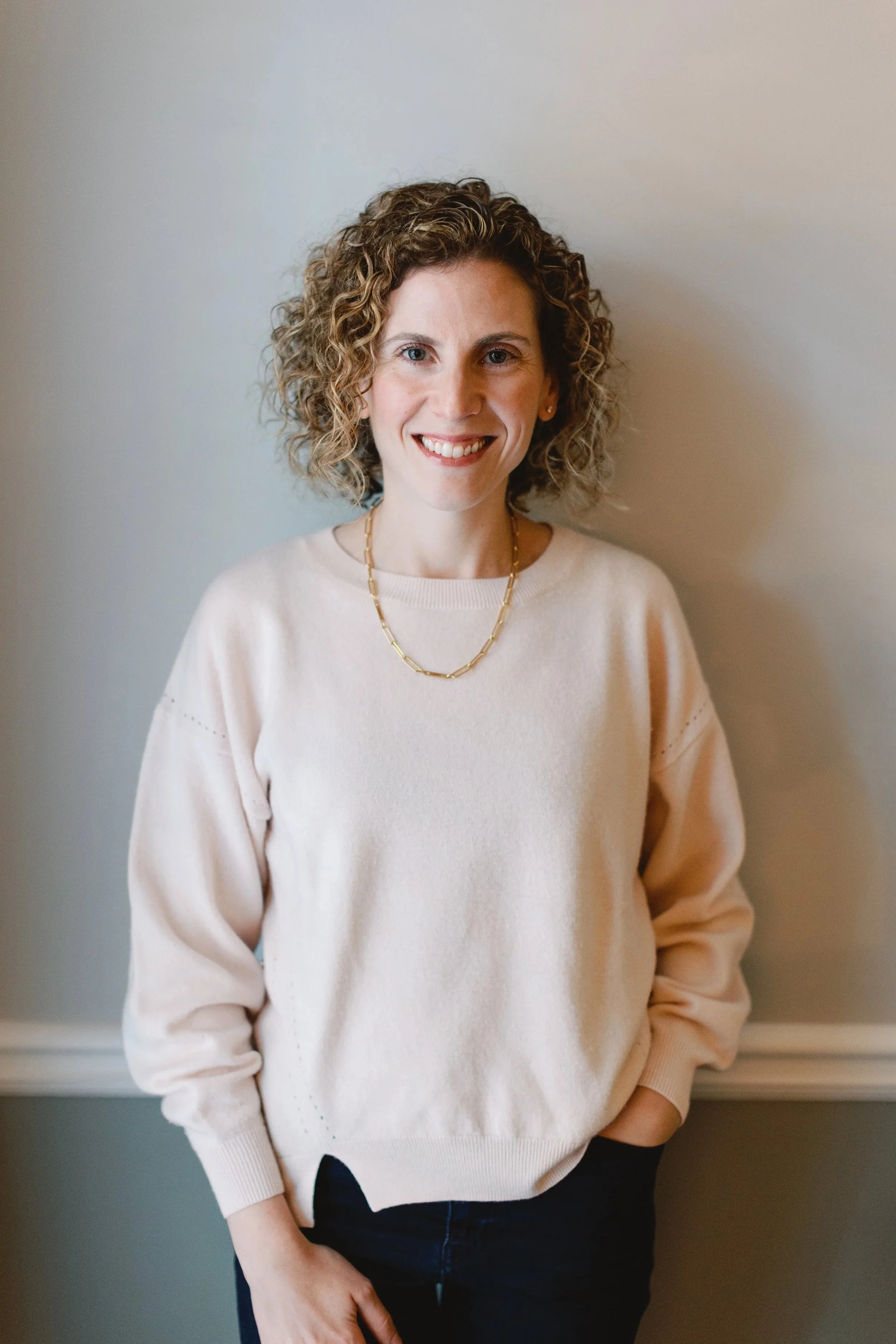 A woman with curly, shoulder-length hair smiling, wearing a light beige sweater and a gold chain necklace, standing in front of a neutral wall. Psychiatric Care - Amy Zoller Lee - PA-C