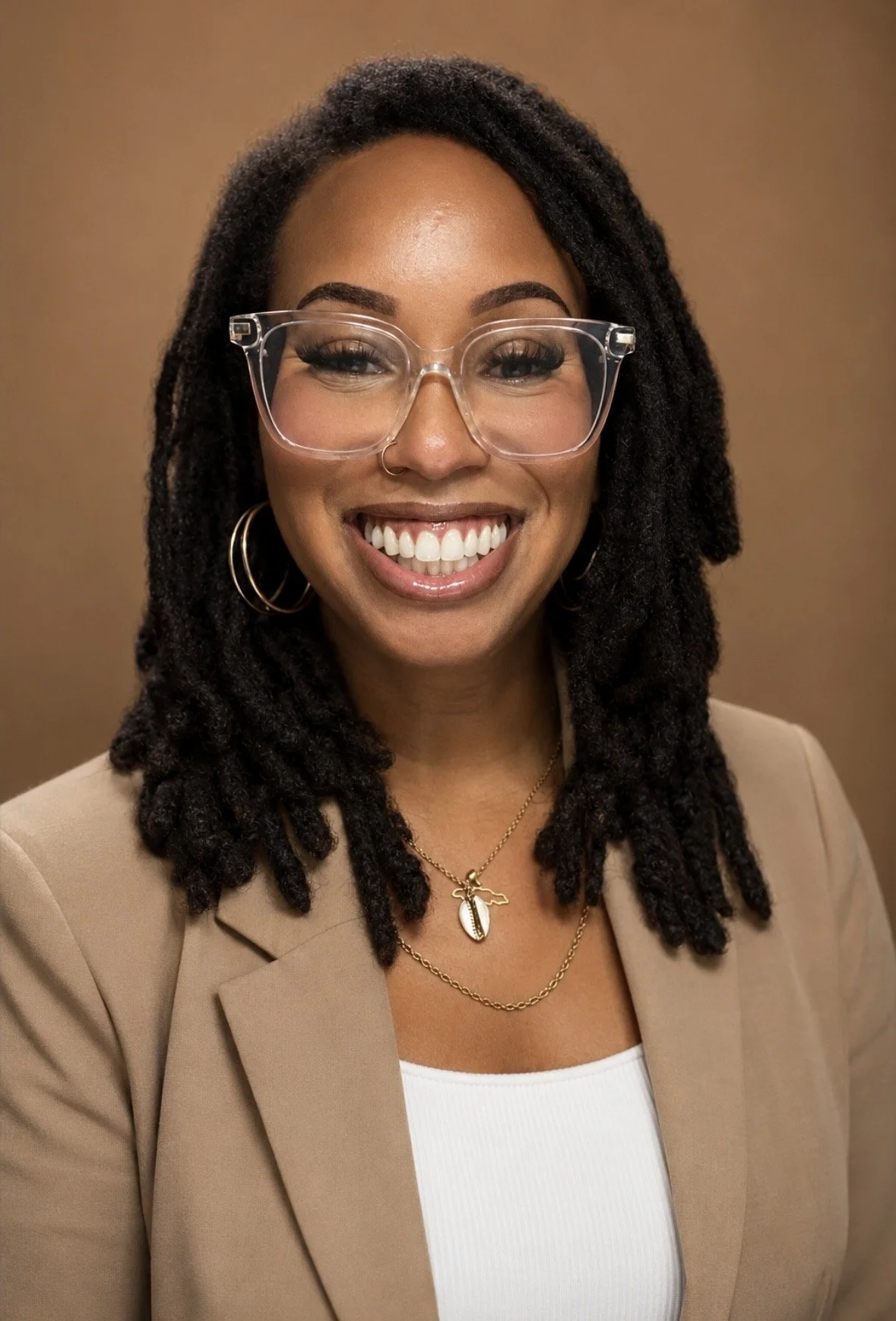 A woman wearing clear glasses, hoop earrings, a beige blazer, and layered necklaces with a smile, against a plain brown background.