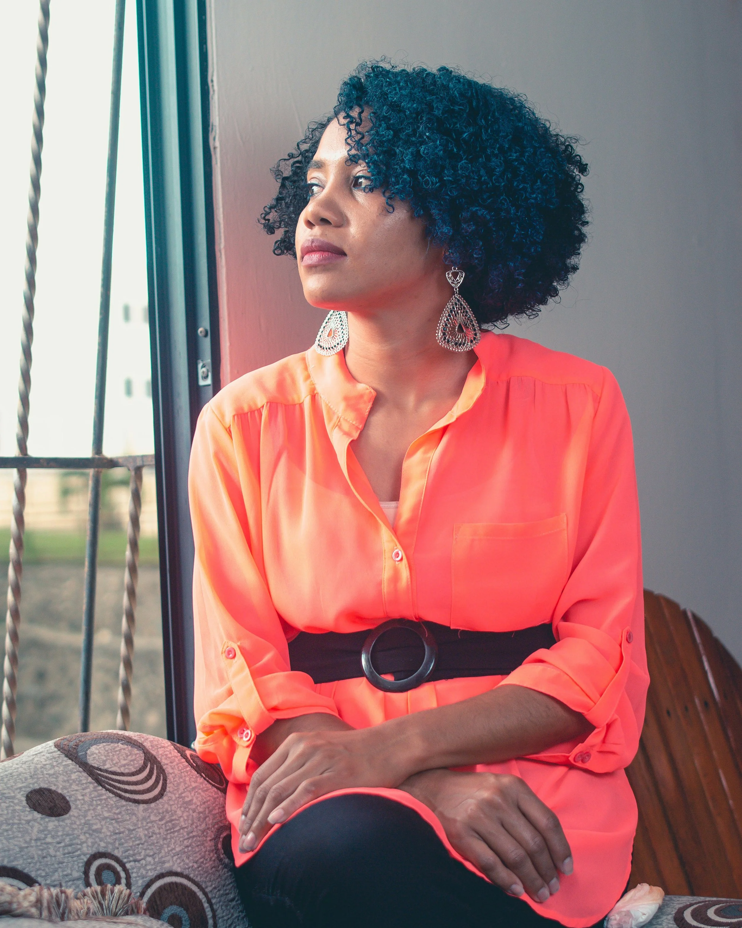 A woman with dark, curly hair and medium skin tone, wearing a bright orange blouse with rolled-up sleeves and large silver earrings, is sitting indoors near a window, looking thoughtfully out of the frame.