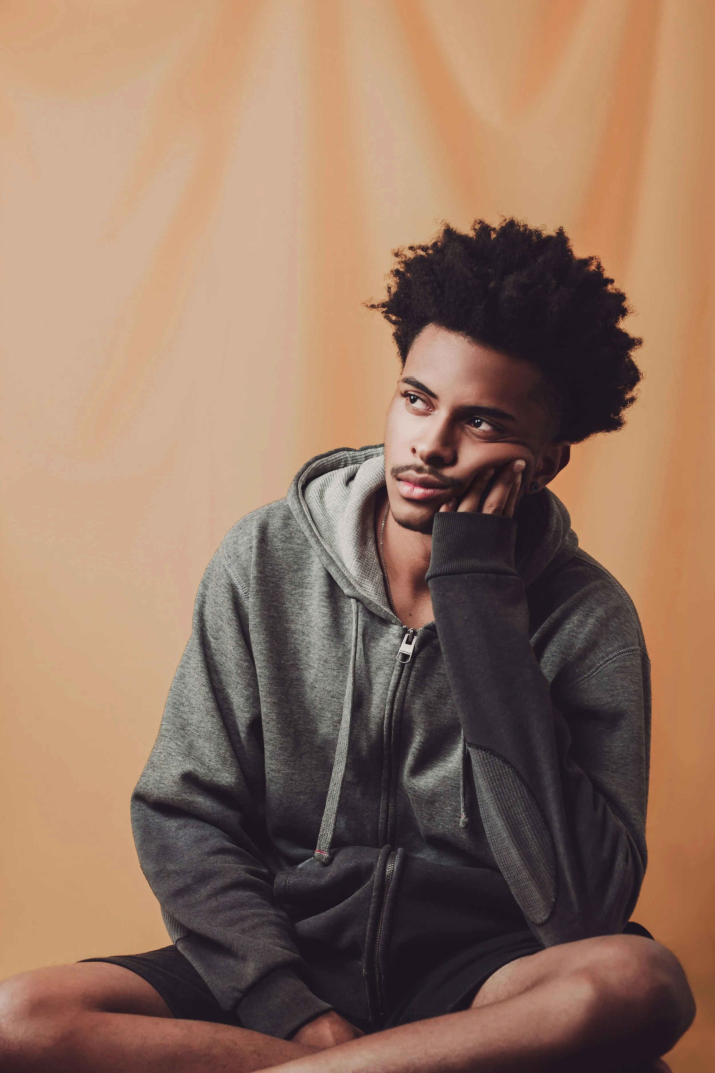A young man with dark, curly hair sitting cross-legged against a beige background. He is wearing a gray hoodie and black shorts, with his left hand resting on his cheek, gazing thoughtfully to the right.