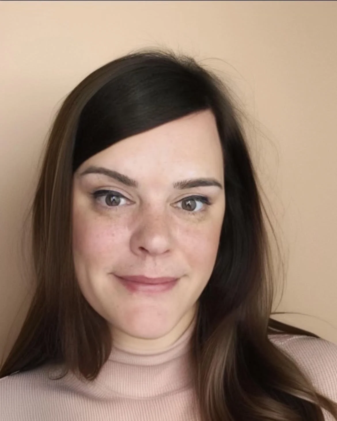 Close-up of a woman with brown hair, wearing a light pink top, standing against a beige wall.