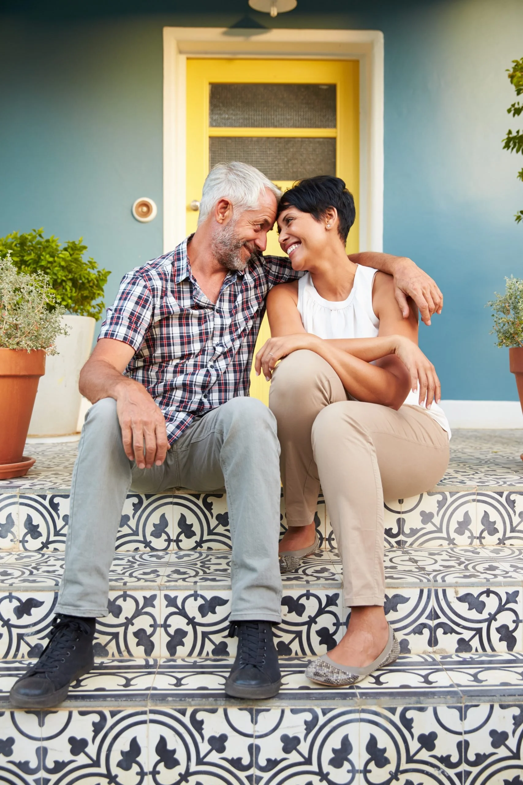 A middle-aged man and woman sitting on decorative tiled steps outside a house, touching foreheads and smiling at each other, with plants on either side.