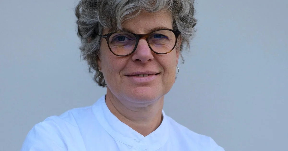 A middle-aged woman with short, curly gray hair and glasses poses against a plain background, wearing a white collared shirt.