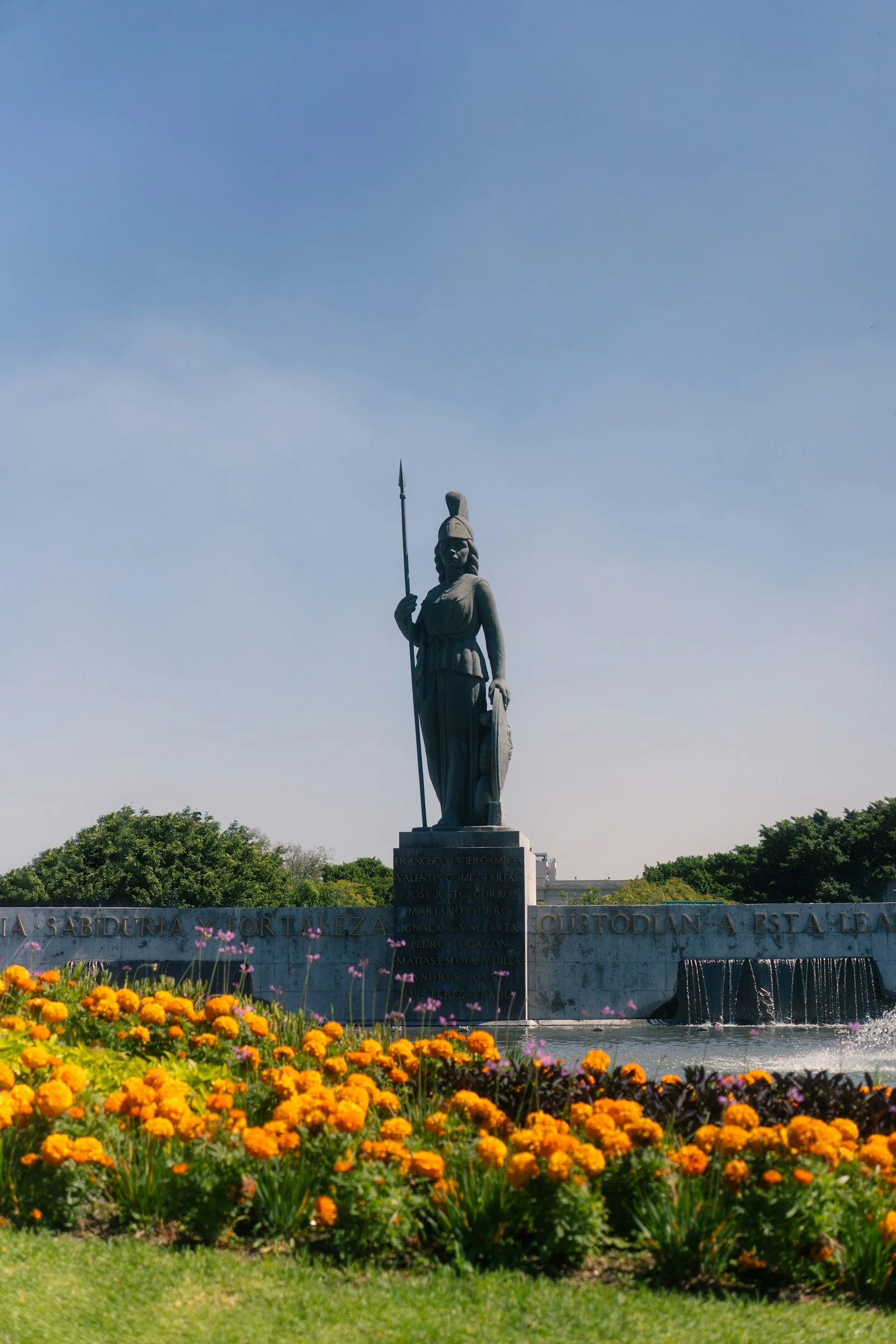 A tall statue of a woman holding a spear and shield, standing on a pedestal with names inscribed. The statue is surrounded by a fountain and colorful orange and pink flowers, with green bushes and trees in the background under a partly cloudy sky.