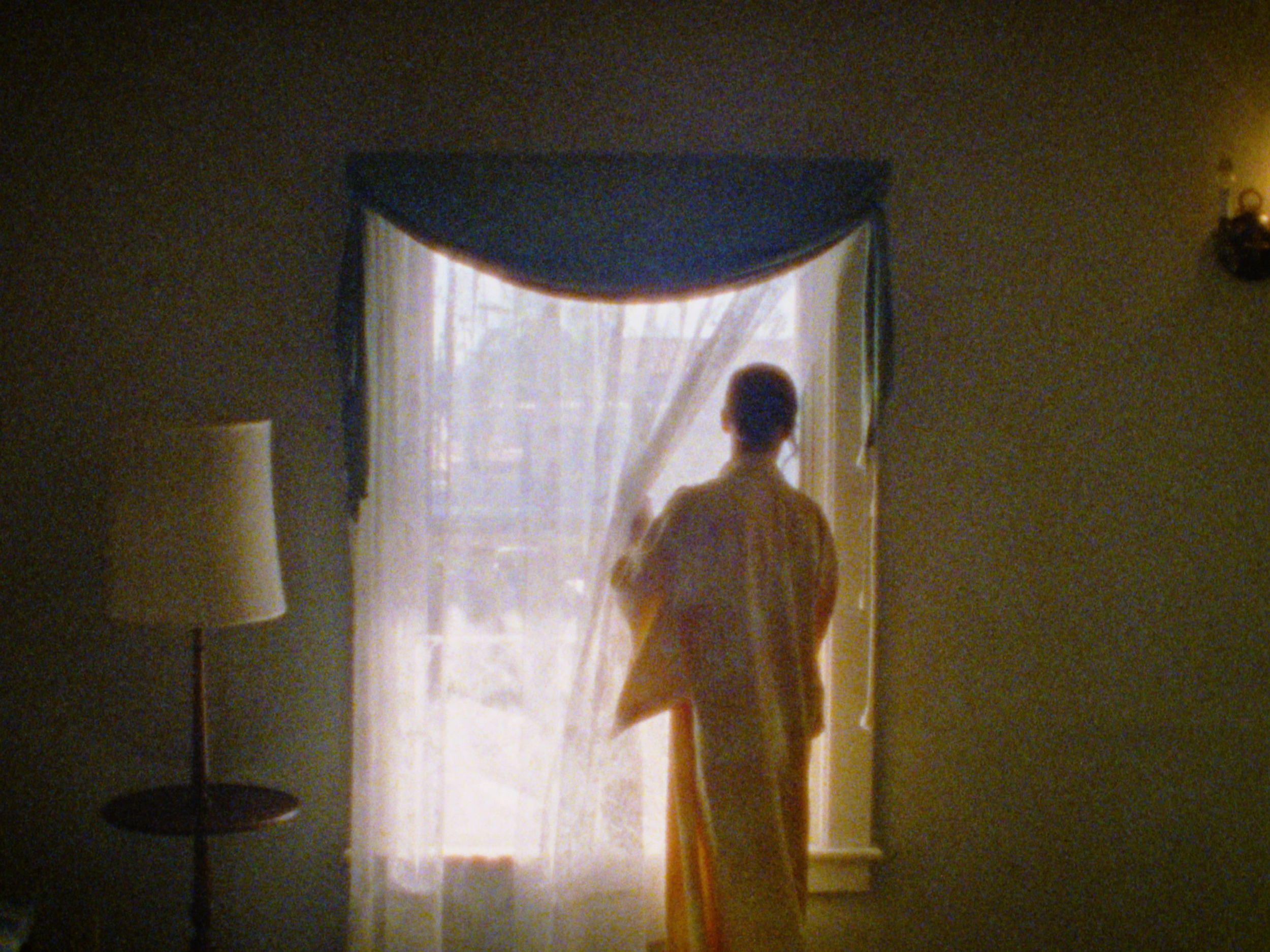 Person standing in front of a window with sheer curtains during daytime, back view, warm lighting, with a floor lamp and wall-mounted light fixture in a home interior.