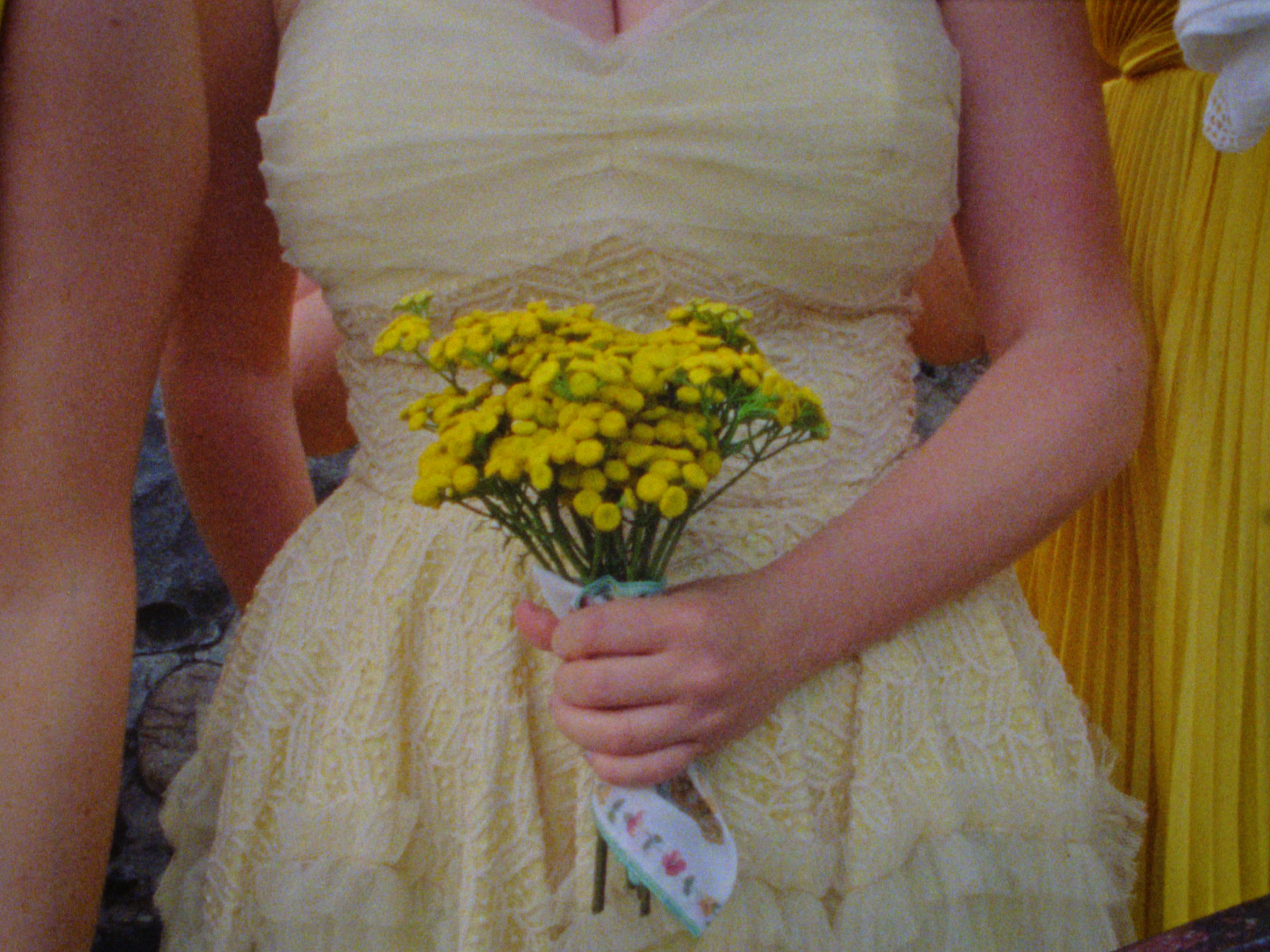 A girl wearing a yellow dress is holding a bouquet of small yellow flowers wrapped in white floral paper.