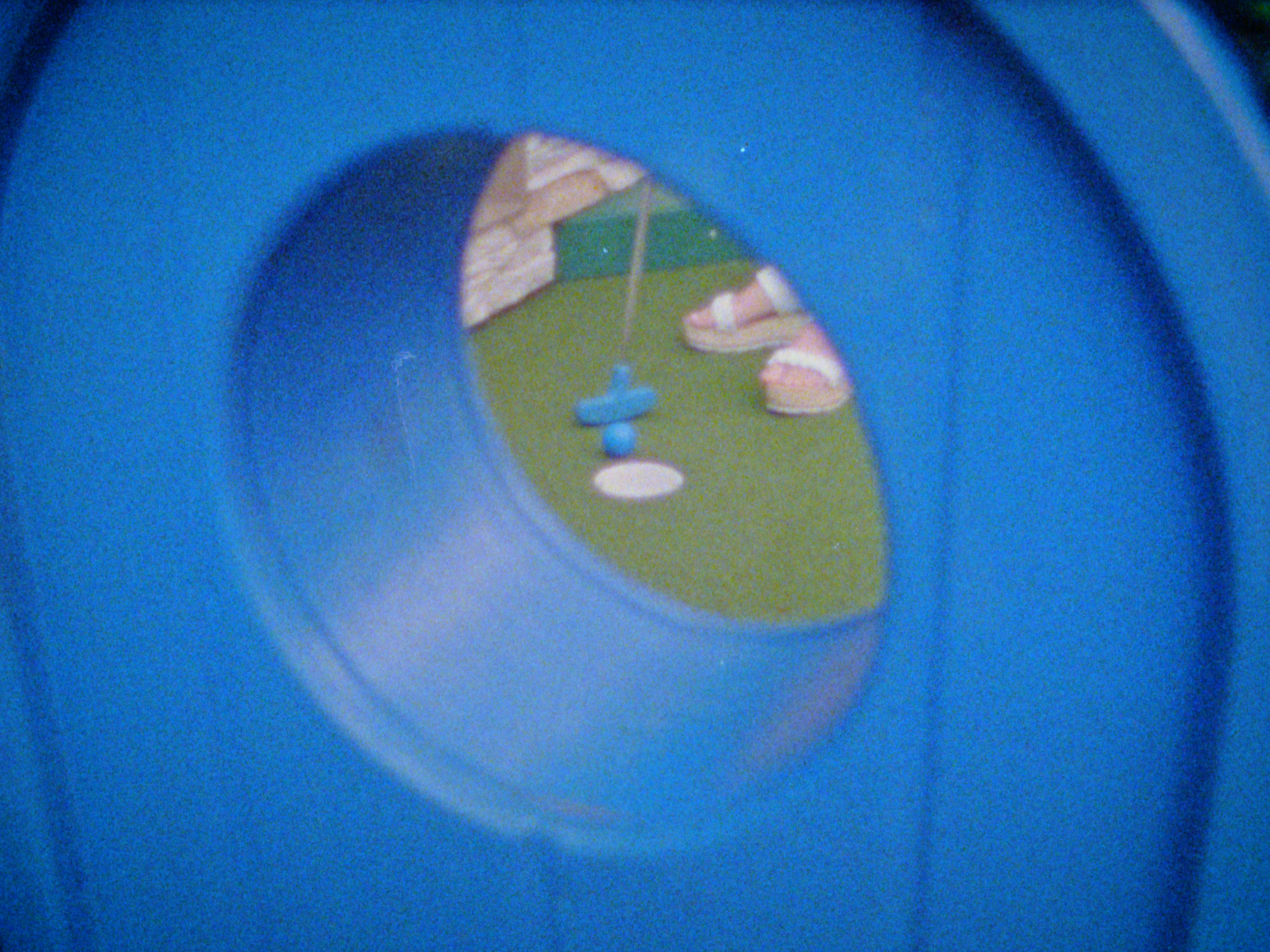 A view through a blue circular tunnel or tube with a small outdoor scene visible at the end, including a structure and some objects on the ground.