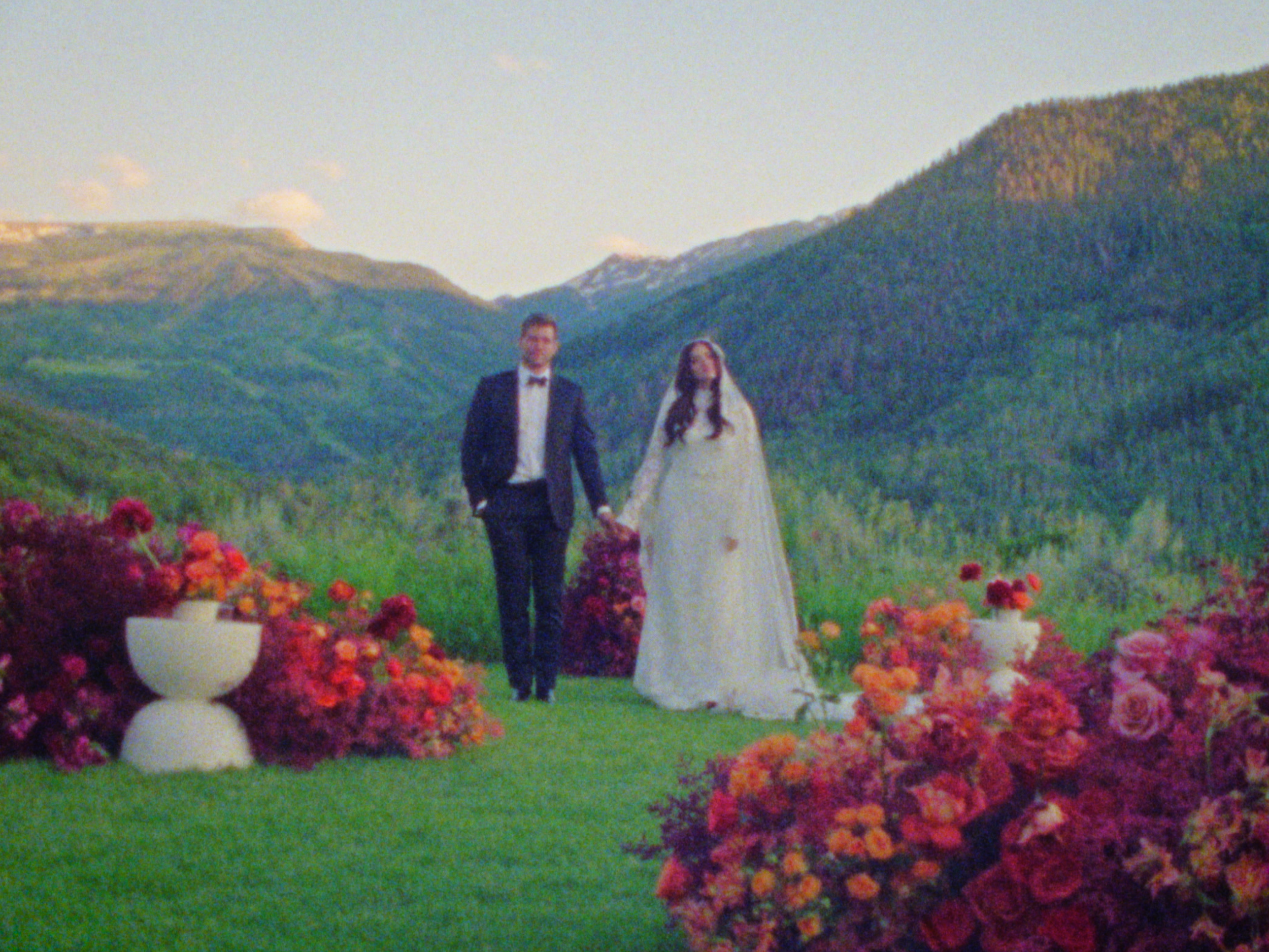 A bride and groom holding hands walking through a lush, green landscape with mountains in the background and colorful flowers in the foreground.