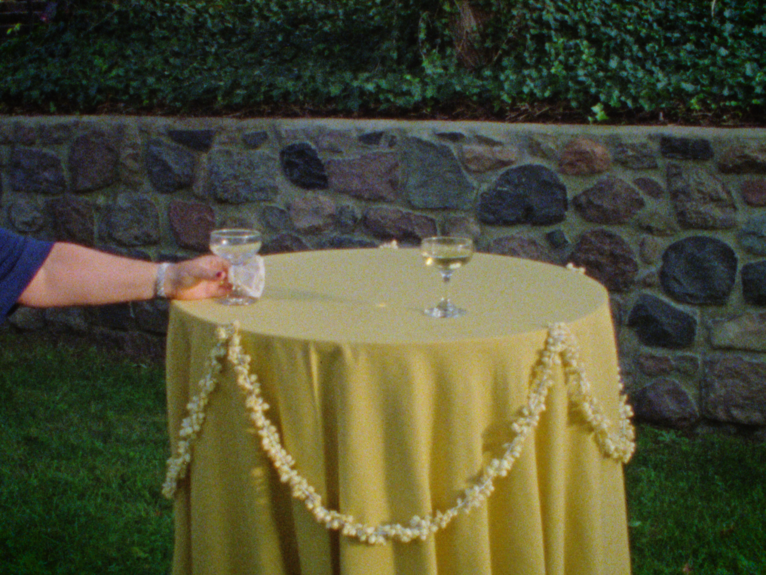 A yellow table with two glasses of white wine, a floral garland, and a stone wall with greenery above, in an outdoor setting.