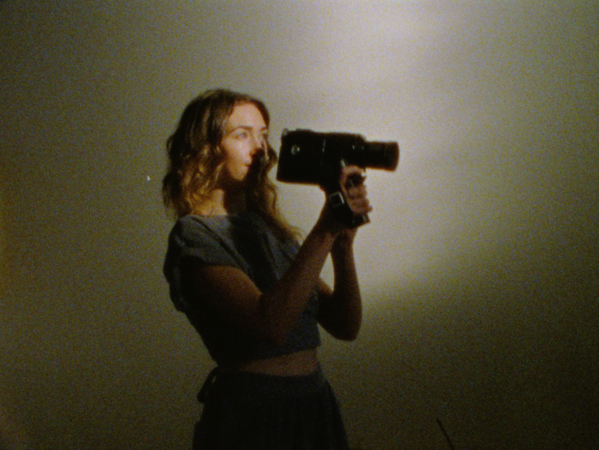 A woman with wavy brown hair takes a mirror selfie with a large black phone against a plain wall.