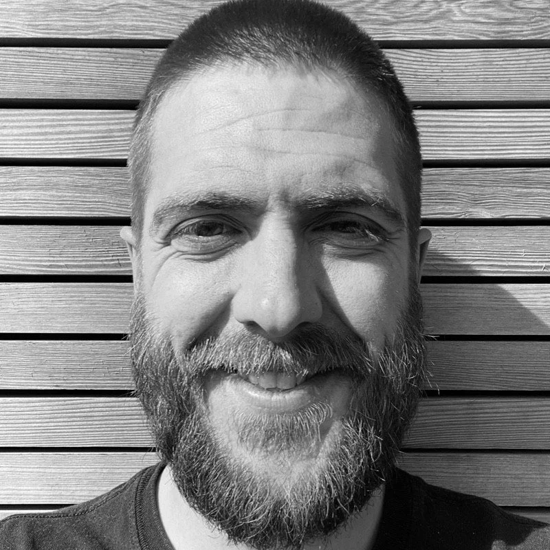 A man with a short beard and mustache smiling outdoors in front of a wooden slat wall, backlit by sunlight.
