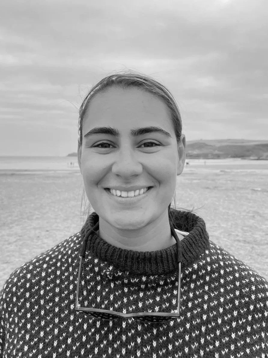 Black and white photo of a smiling woman with sunglasses hanging from her sweater, on a beach with cloudy sky and distant hills.
