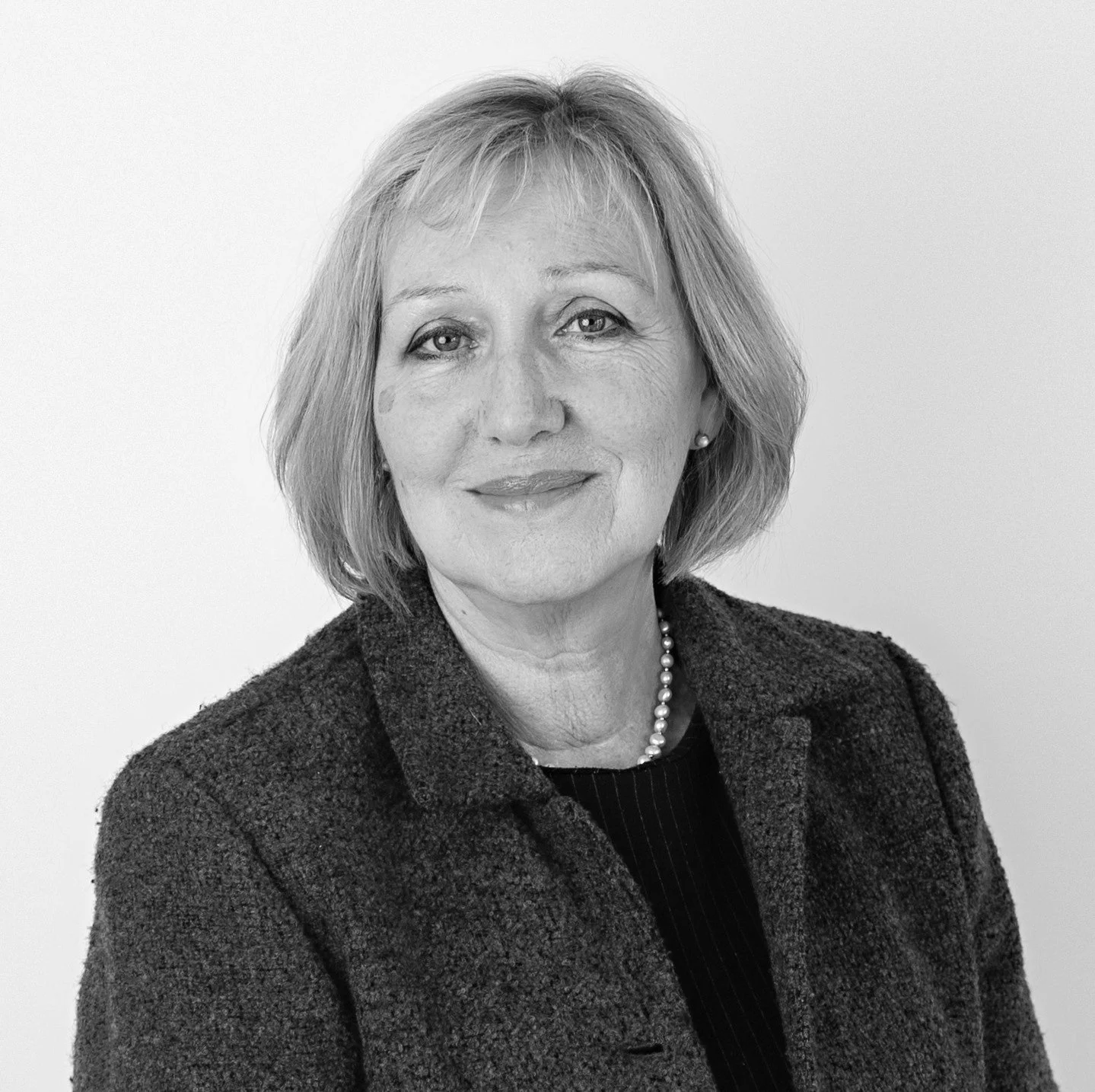 Black and white portrait of a middle-aged woman with shoulder-length hair, wearing a dark blazer, a pearl necklace, and a dark top, standing against a plain light background, smiling slightly.