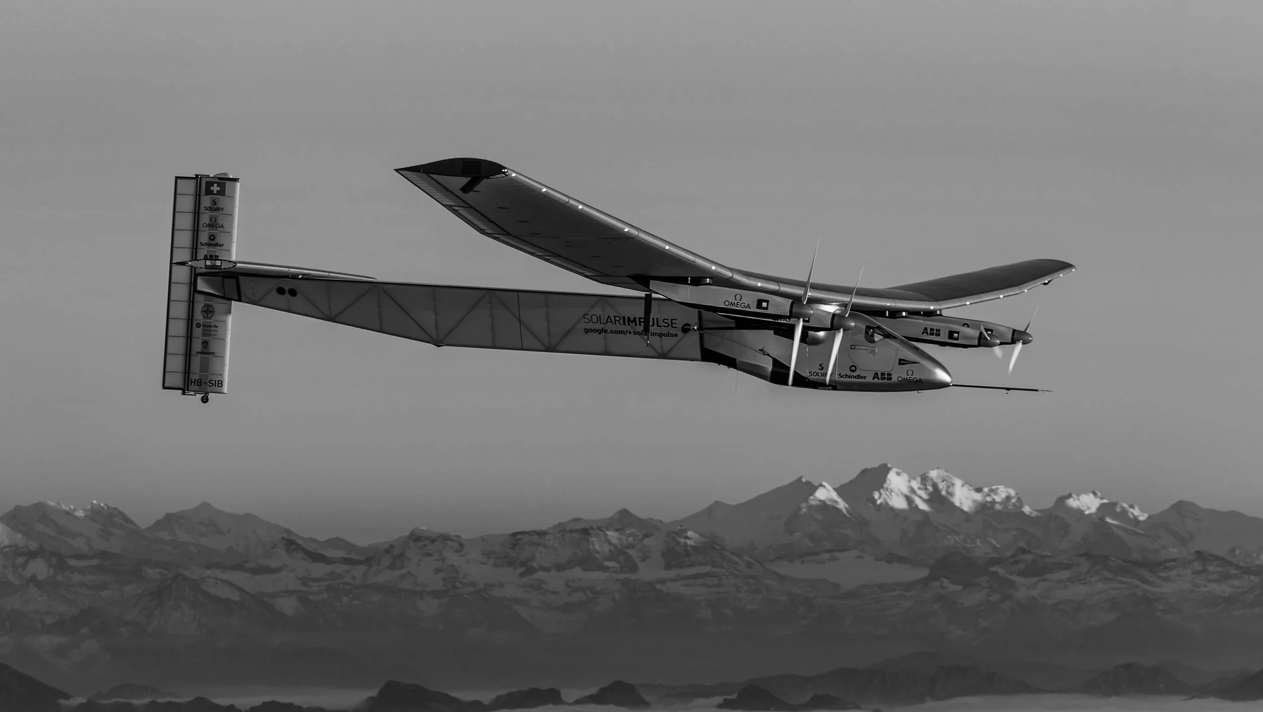 A solar-powered aircraft flying above snow-capped mountains in a black-and-white photograph.