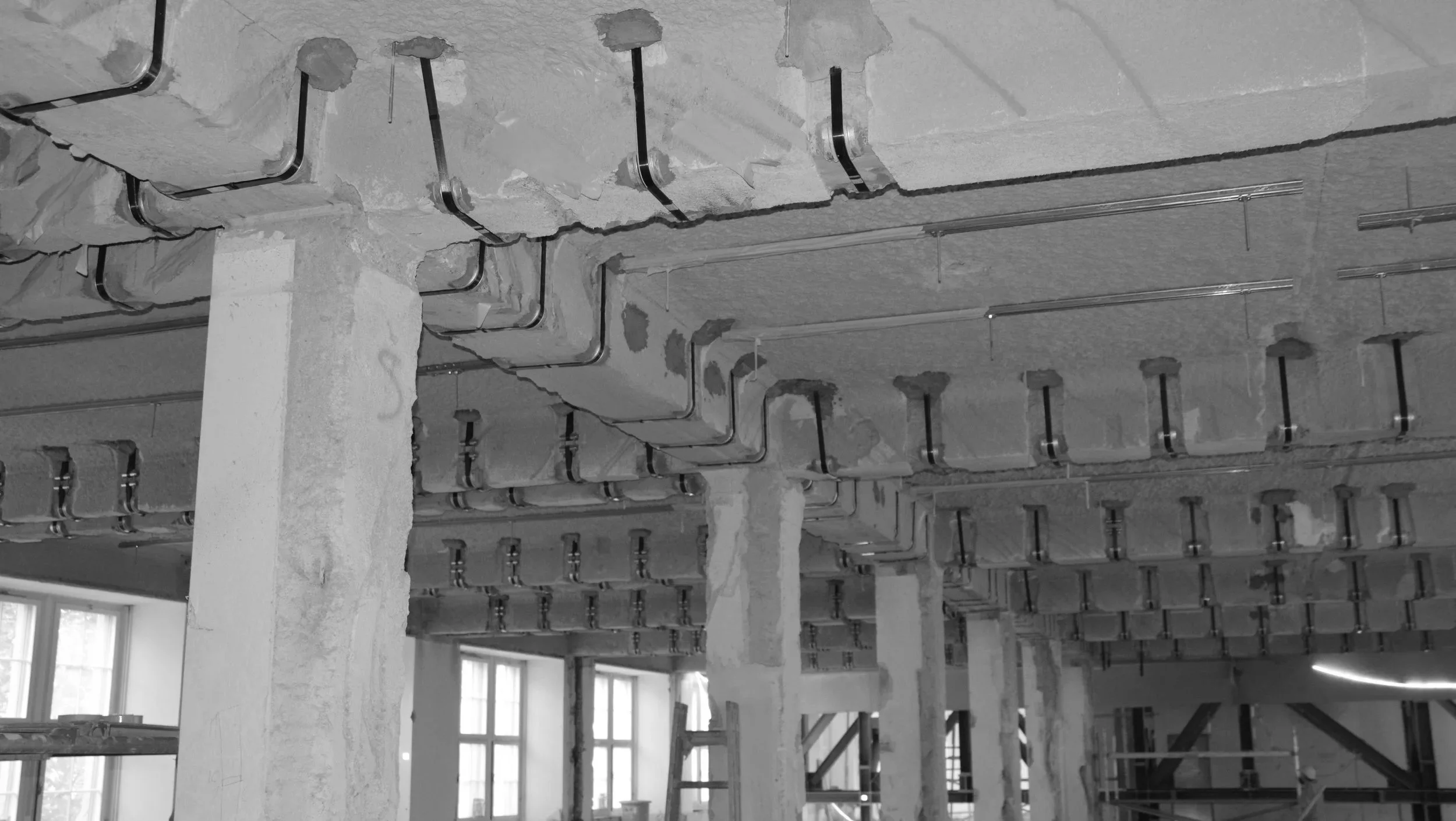 Construction site showing exposed concrete beams with installed vertical and horizontal metal pipes, support scaffolding, and large windows in the background.
