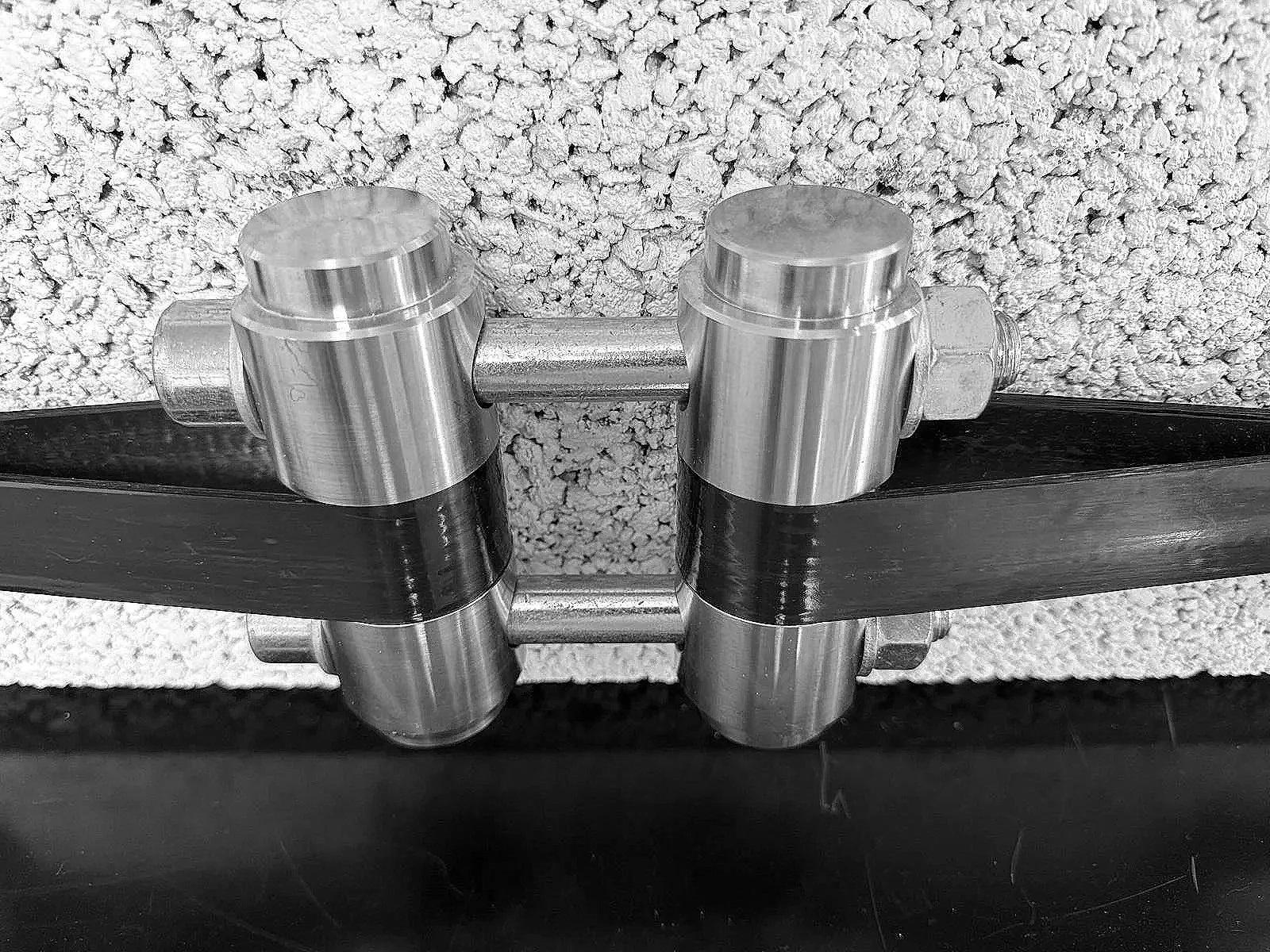 A metal plumbing fitting with two parallel black pipes connected, mounted on a textured wall.