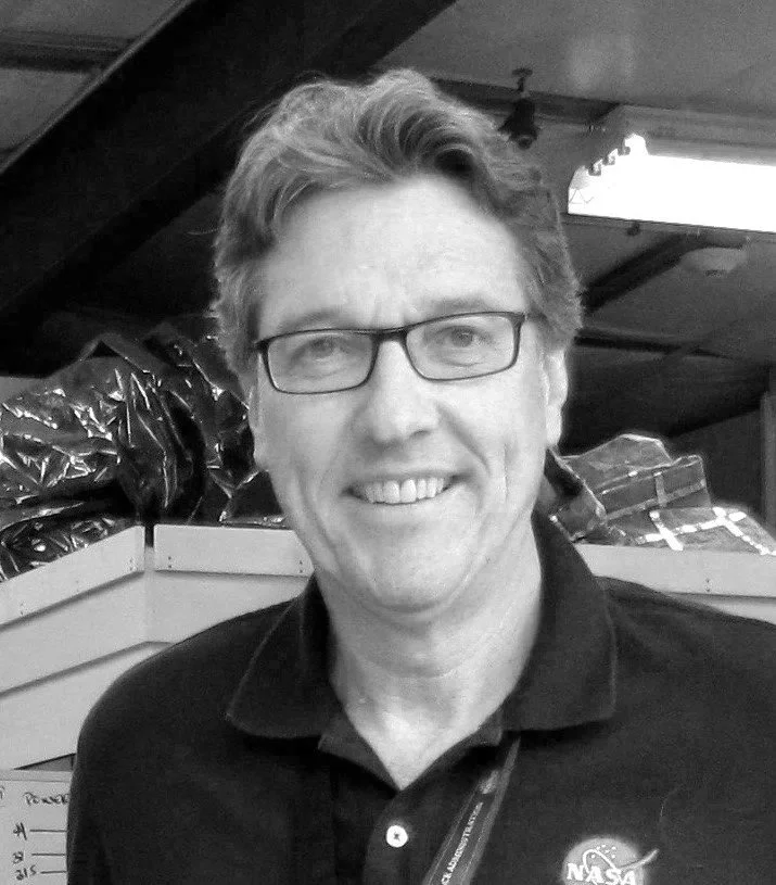 A smiling man with glasses and light-colored hair, wearing a NASA polo shirt, standing indoors with boxes and plastic bags in the background.