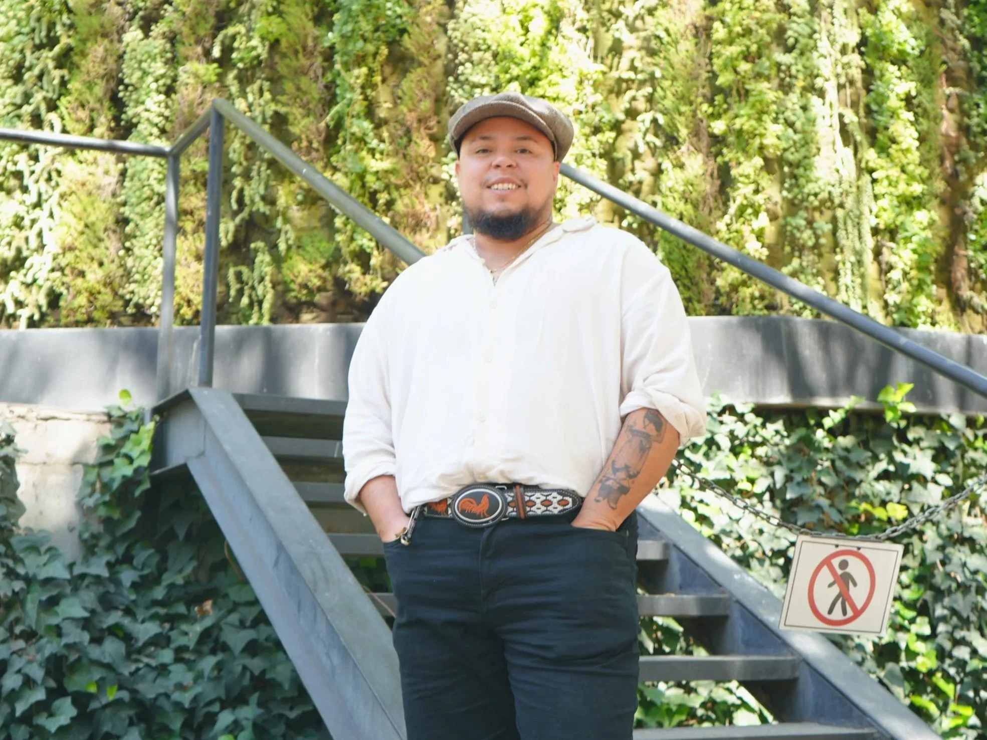 A man with a beard, wearing a light-colored shirt, a hat, and tattoos on one arm, standing on outdoor stairs with greenery behind him.
