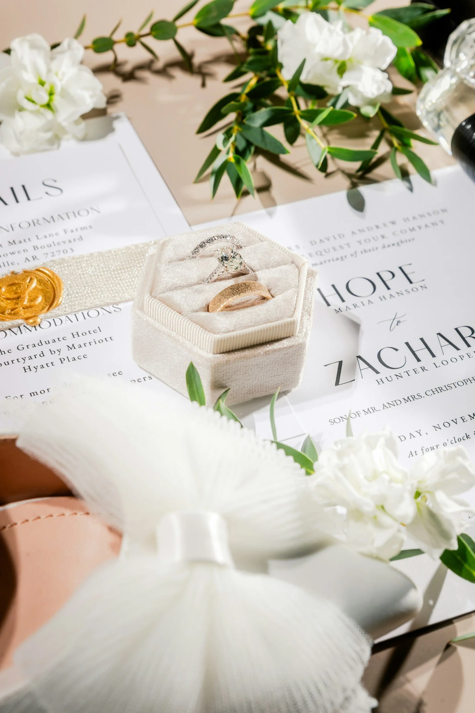 A wedding invitation surrounded by white flowers and greenery, with a box containing a diamond engagement ring and two gold wedding bands placed on it.