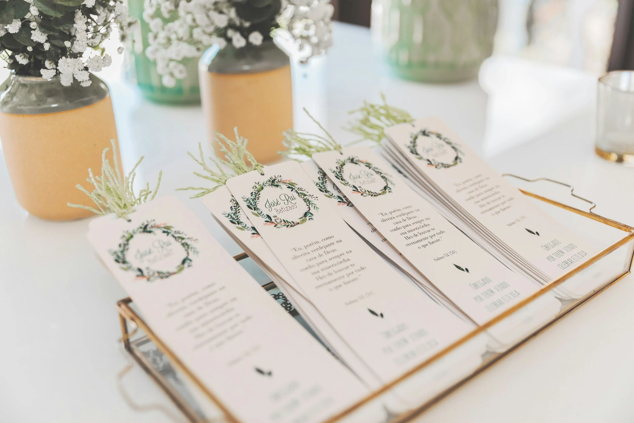 Wedding programs featuring a green wreath with a white background, arranged on a table with floral decorations in vases, including white flowers and greenery.