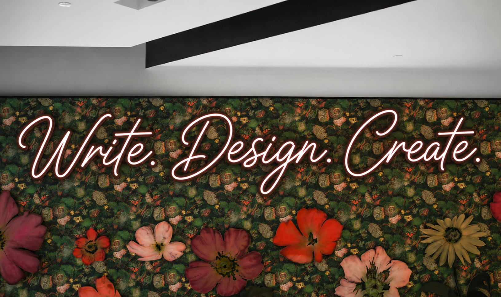 Neon sign that says 'Write. Design. Create.' against a floral background with various colorful flowers.