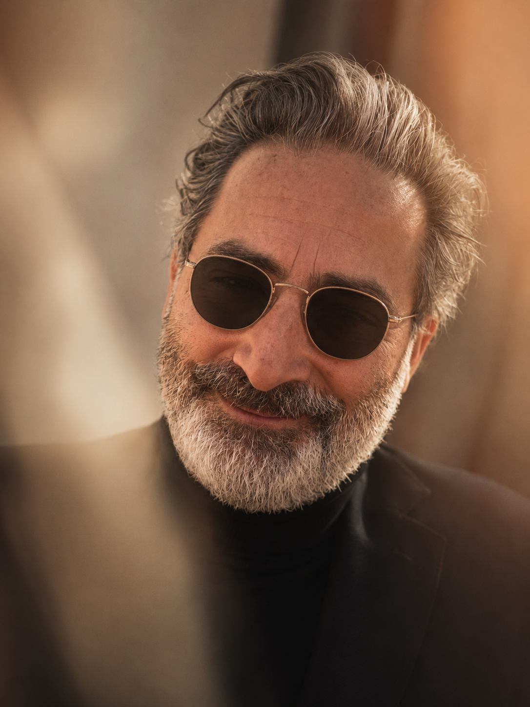 A smiling middle-aged man with gray hair and beard wearing sunglasses and a dark suit.