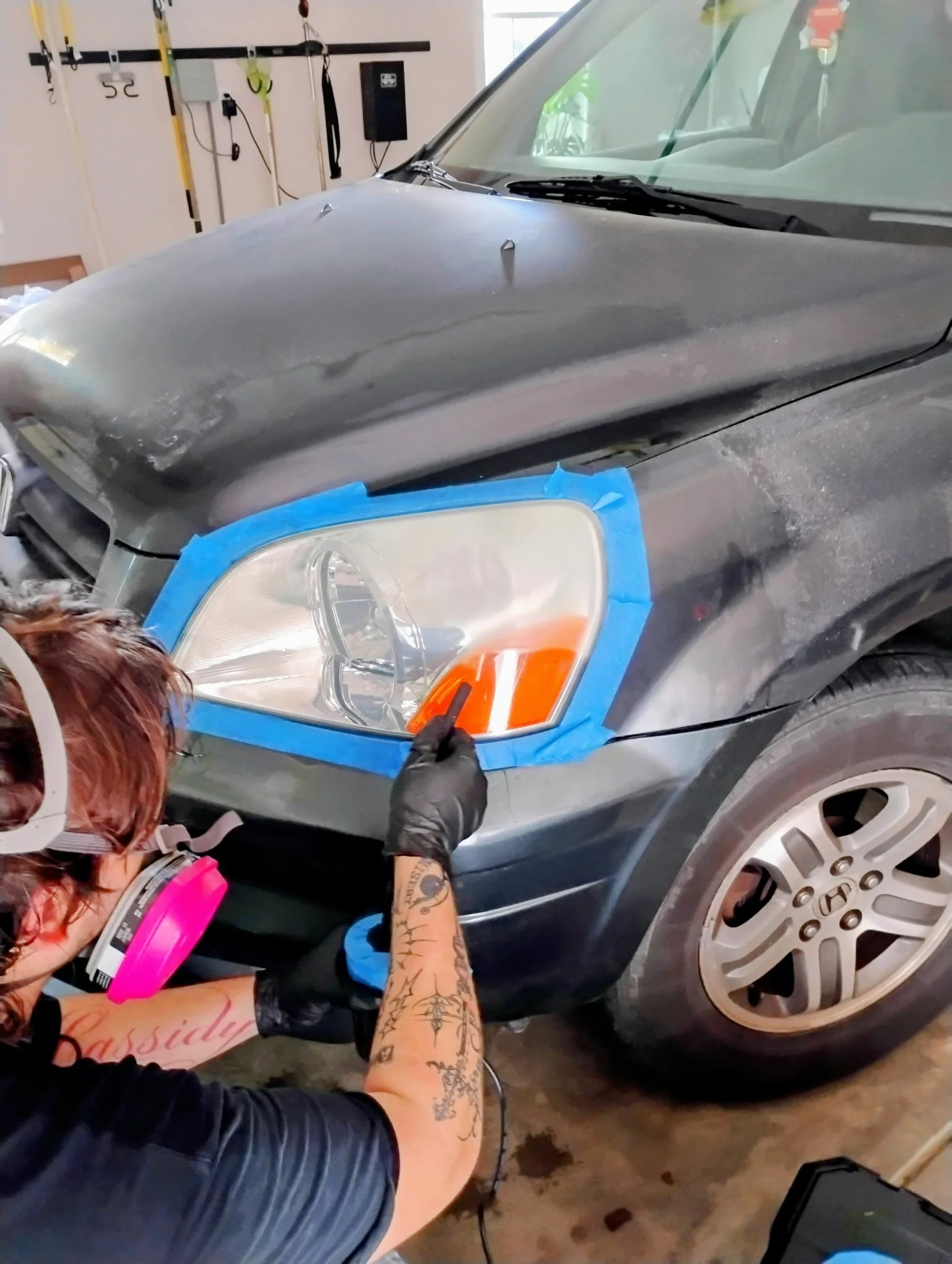 Person working on the headlights of a dark-colored car, which is taped around the headlights, in a garage.