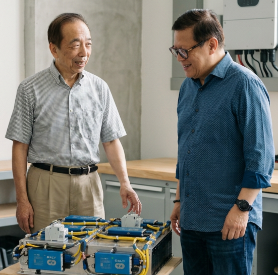 Two men, one older and one younger, are smiling and looking at a technical device or model on a table in an industrial or workshop setting.