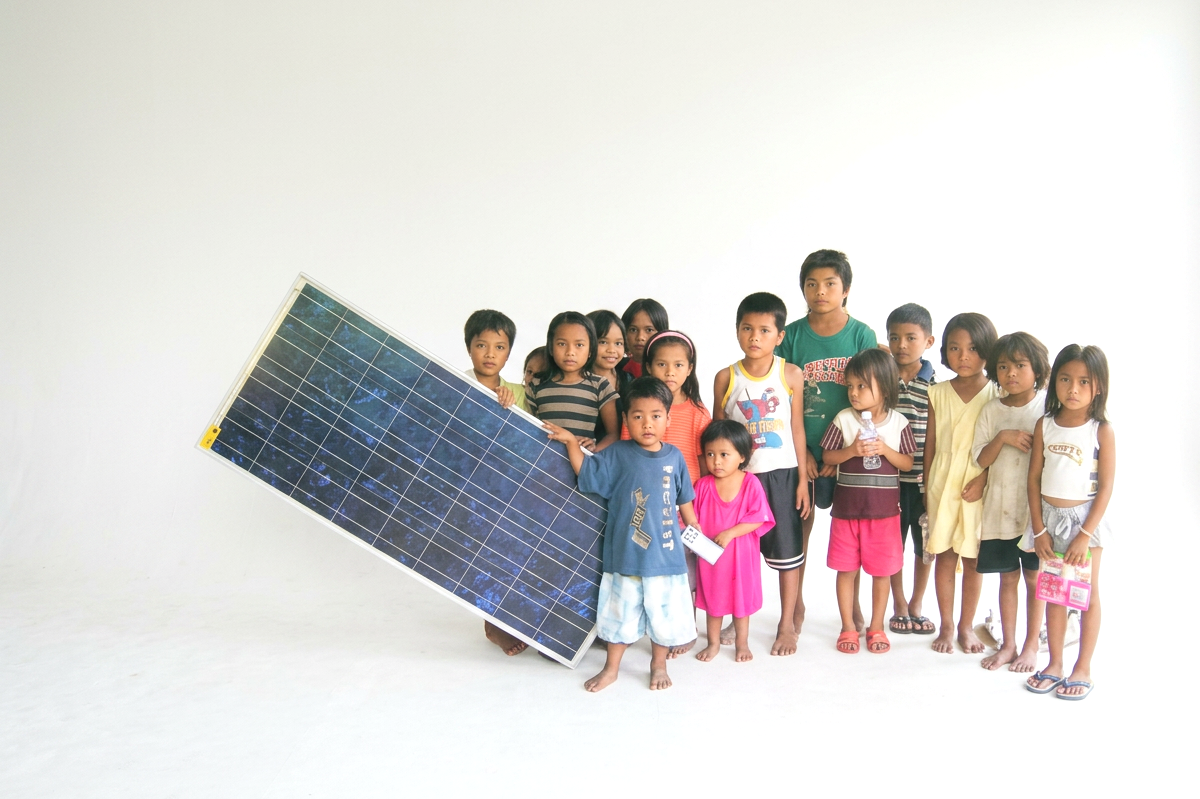 Children of various ages standing together, with one holding a large solar panel, in a bright room with white background.