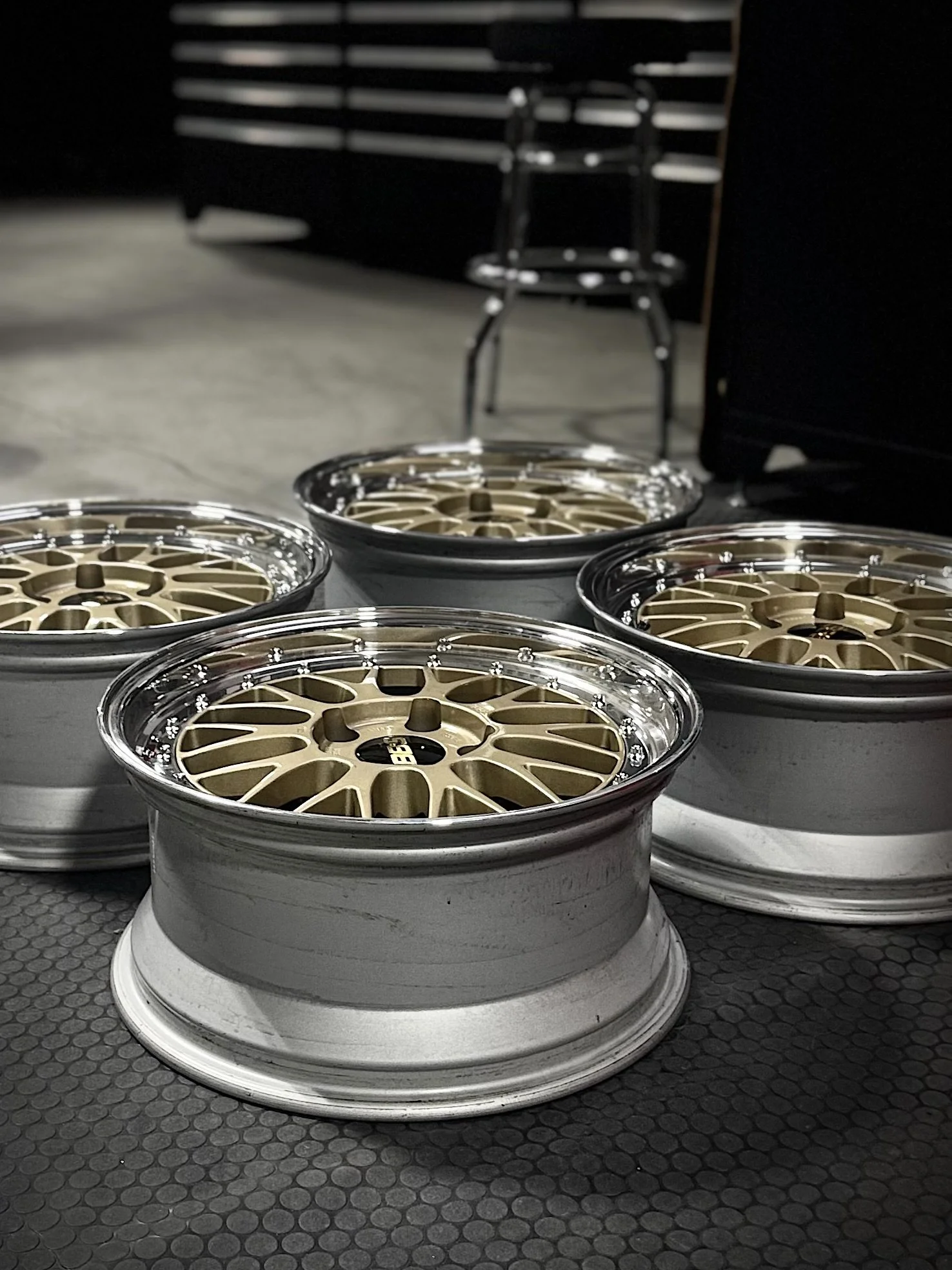 A collection of gold and silver car wheels stacked on the floor in a garage with a black backdrop and slatted wall in the background.