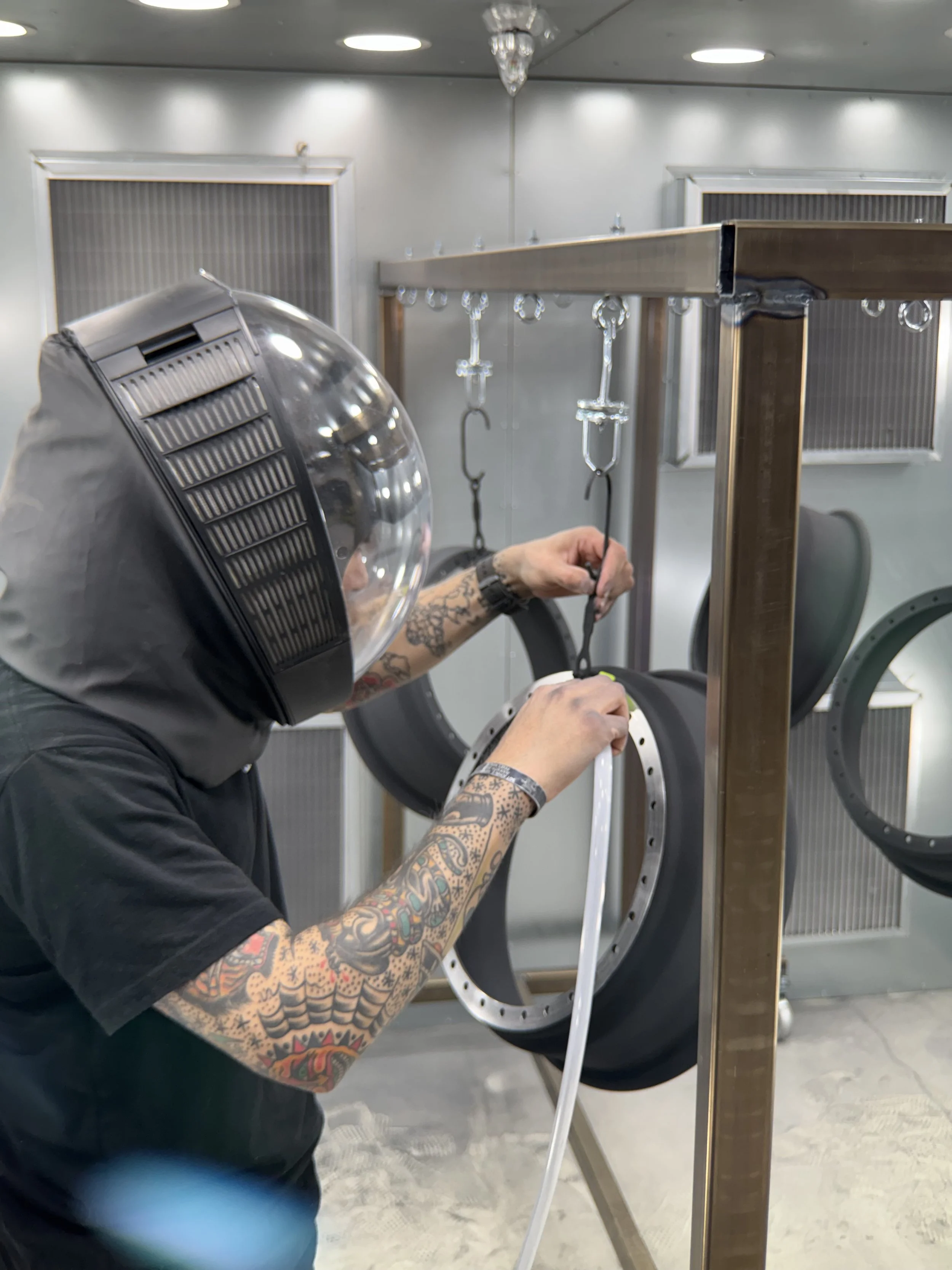 A person with tattooed arms wearing a helmet working on a metal wheel frame, in a room with gray walls, ceiling lights, and windows with blinds, using a tool to assemble or repair the wheel.