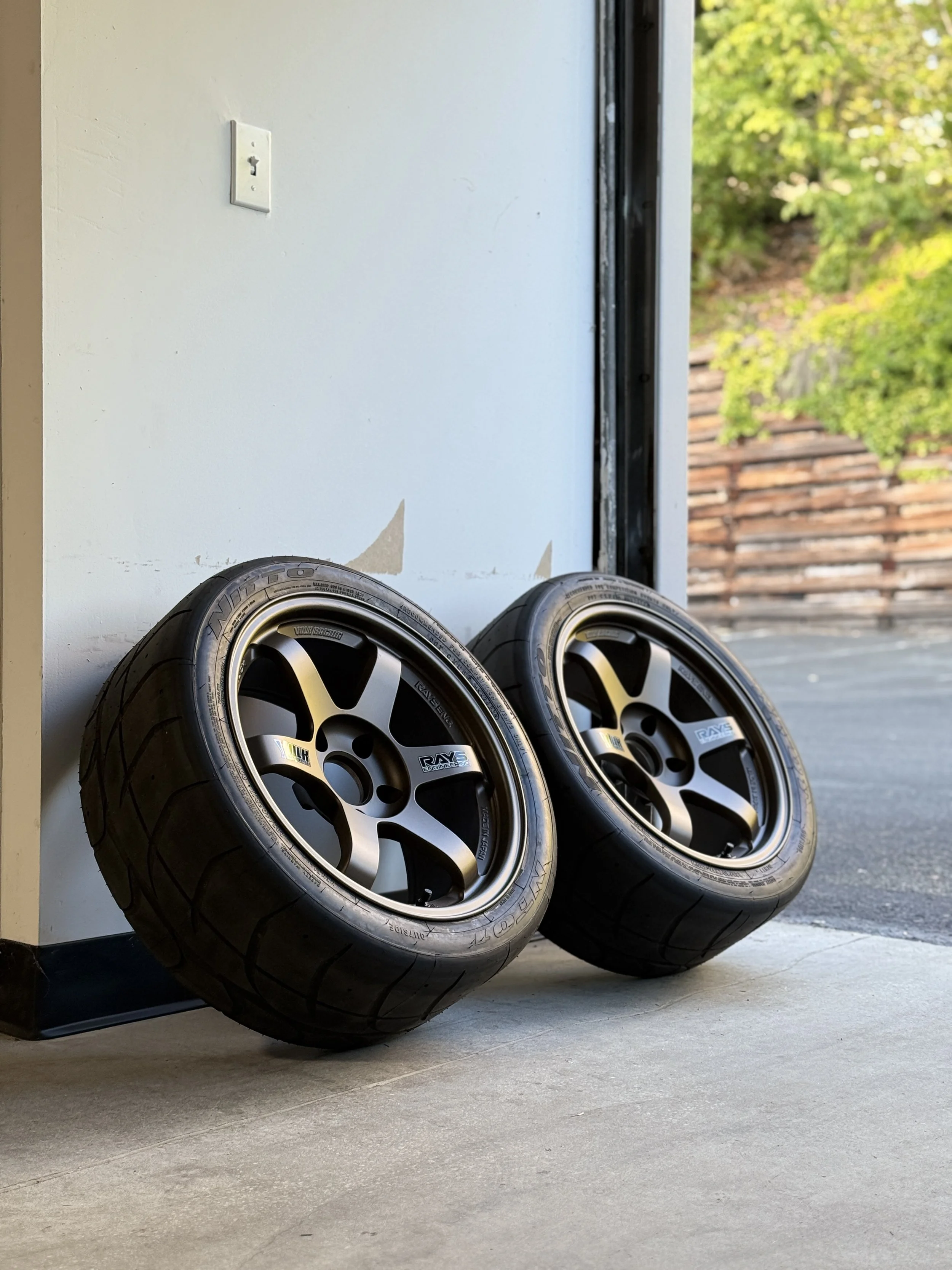 Two racing tires with black rims leaning against a white wall beside a doorway, with a view of trees and a wooden fence outside.