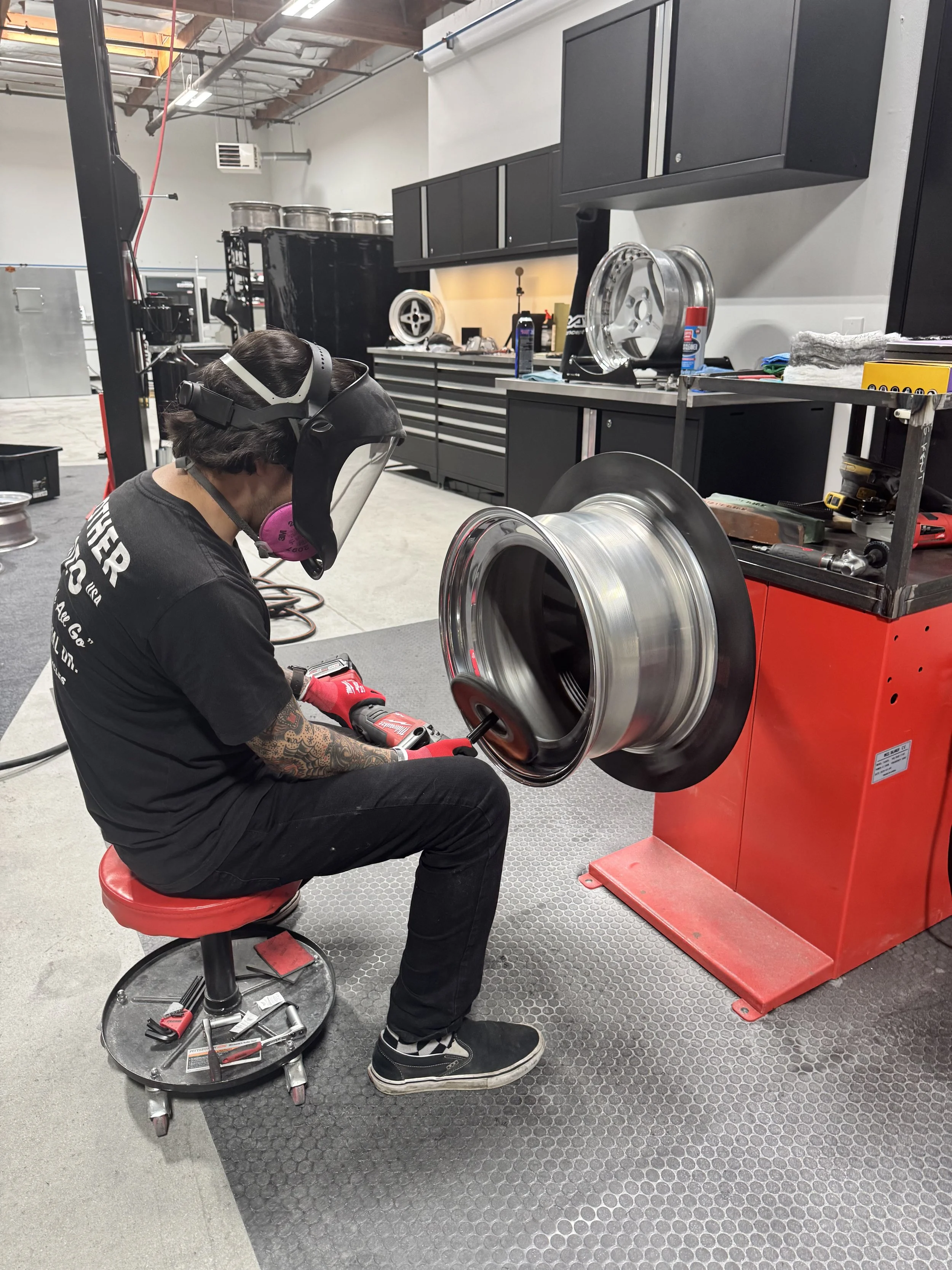 Person wearing a face shield, a mask, and gloves working on a car wheel in a garage or workshop. The person is sitting on a red stool, using tools from a tray on a wheeled stand. The background features workbenches, shelves, tools, and equipment.