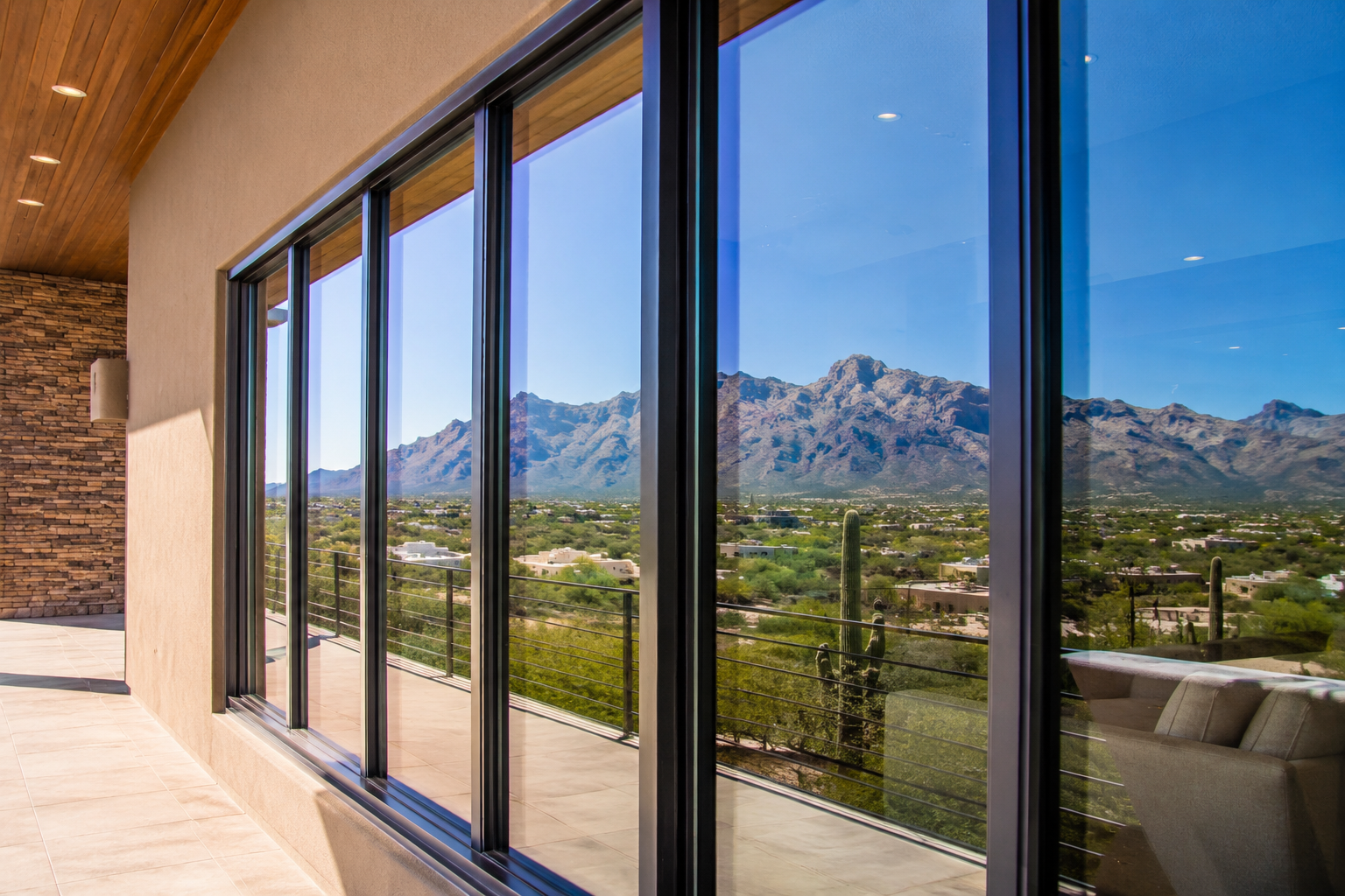 Room with large glass windows revealing mountain and desert landscape outside, including cacti, and interior with beige walls, tiled floor, and sofa.