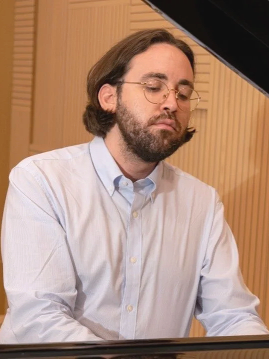 Man with glasses and beard playing a grand piano in a room with wood paneling.