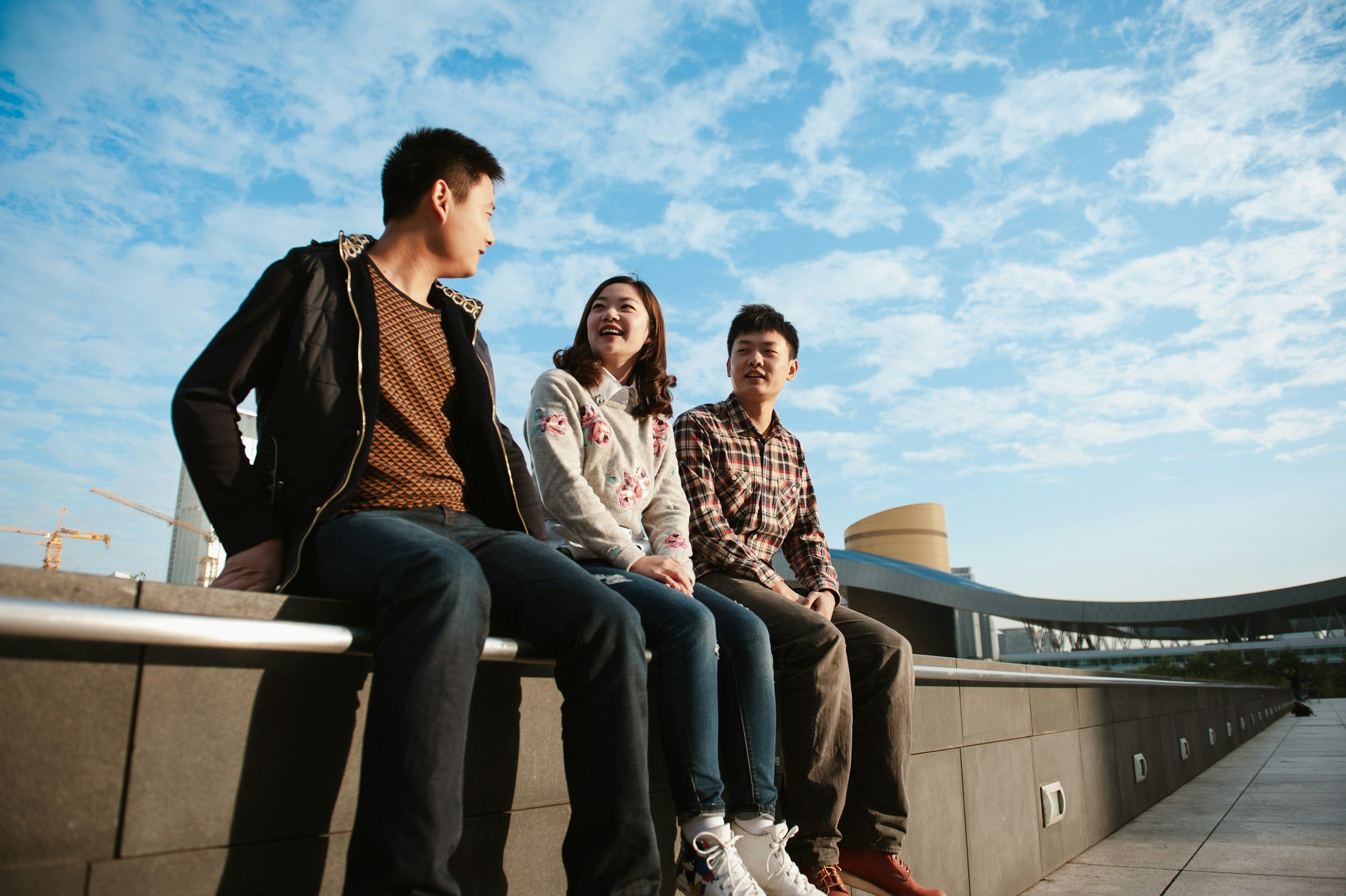 Three young people sitting on a ledge outdoors, engaging in conversation against a background of a building under construction and a blue sky with scattered clouds.