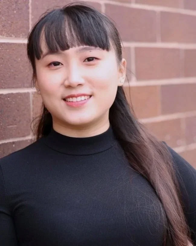 A young woman with dark hair and bangs smiling, wearing a black turtleneck sweater, standing against a brick wall background.