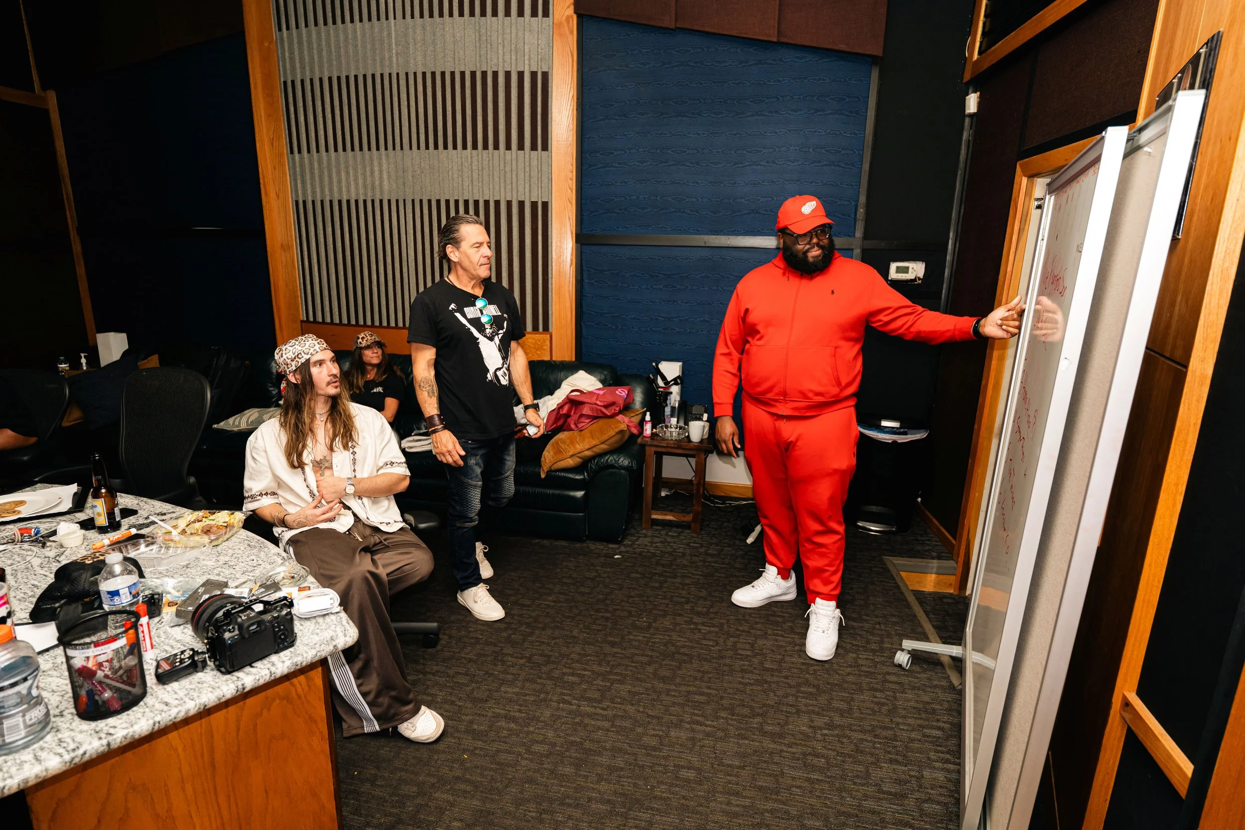 A man in a red hoodie and matching red cap standing and writing on a whiteboard, while three other people sit and stand nearby in a room with blue and wooden walls. The table in foreground has plates, drinks, and camera equipment.