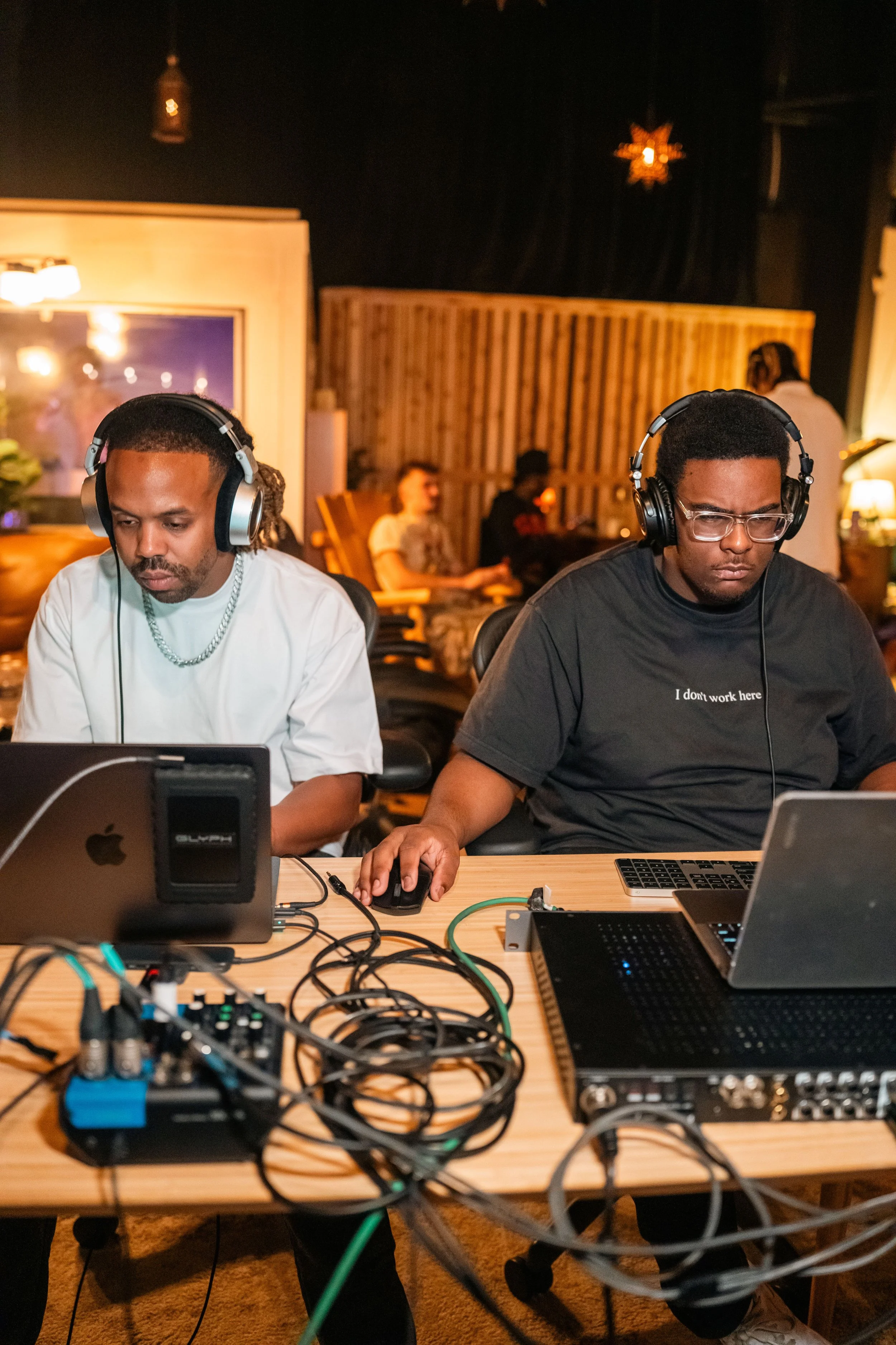 Two men sit at a desk in a recording studio wearing headphones, with computers and audio equipment in front of them, working on a music project.
