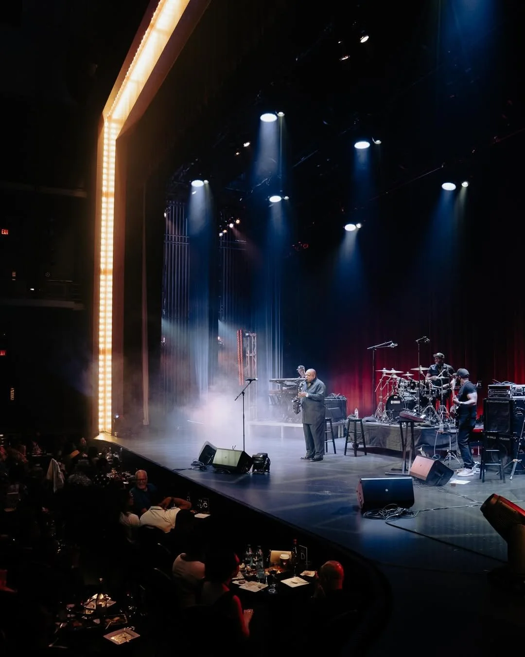 Jazz band performing on stage in a theater, with a saxophonist, drums, keyboard, and microphone, under blue and white lighting.