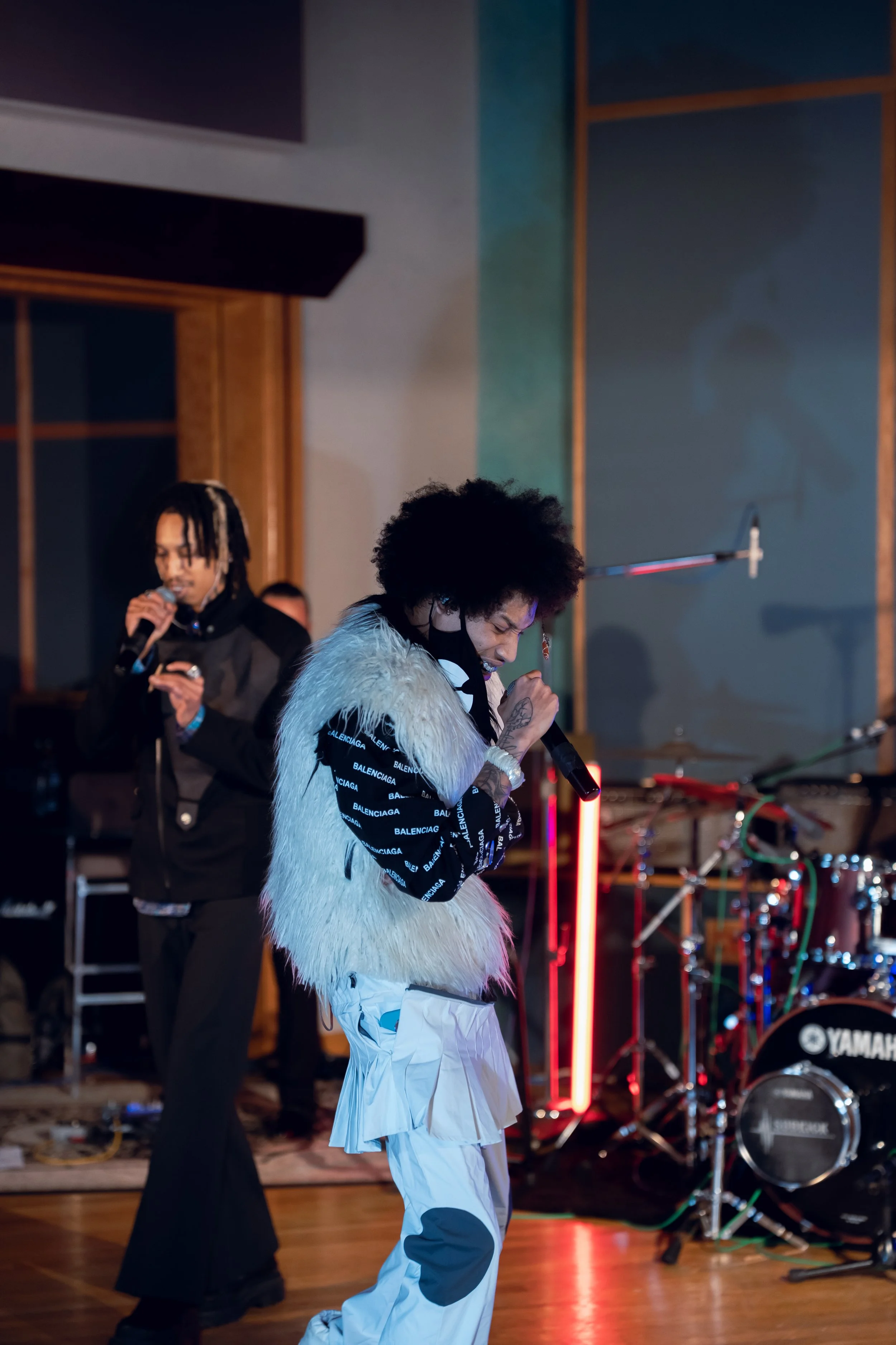 Two musicians performing on stage, one singing into a microphone wearing a white furry vest and white pants with black patches, the other singing with a microphone and wearing a black Balenciaga jacket, with musical instruments and colorful lights in