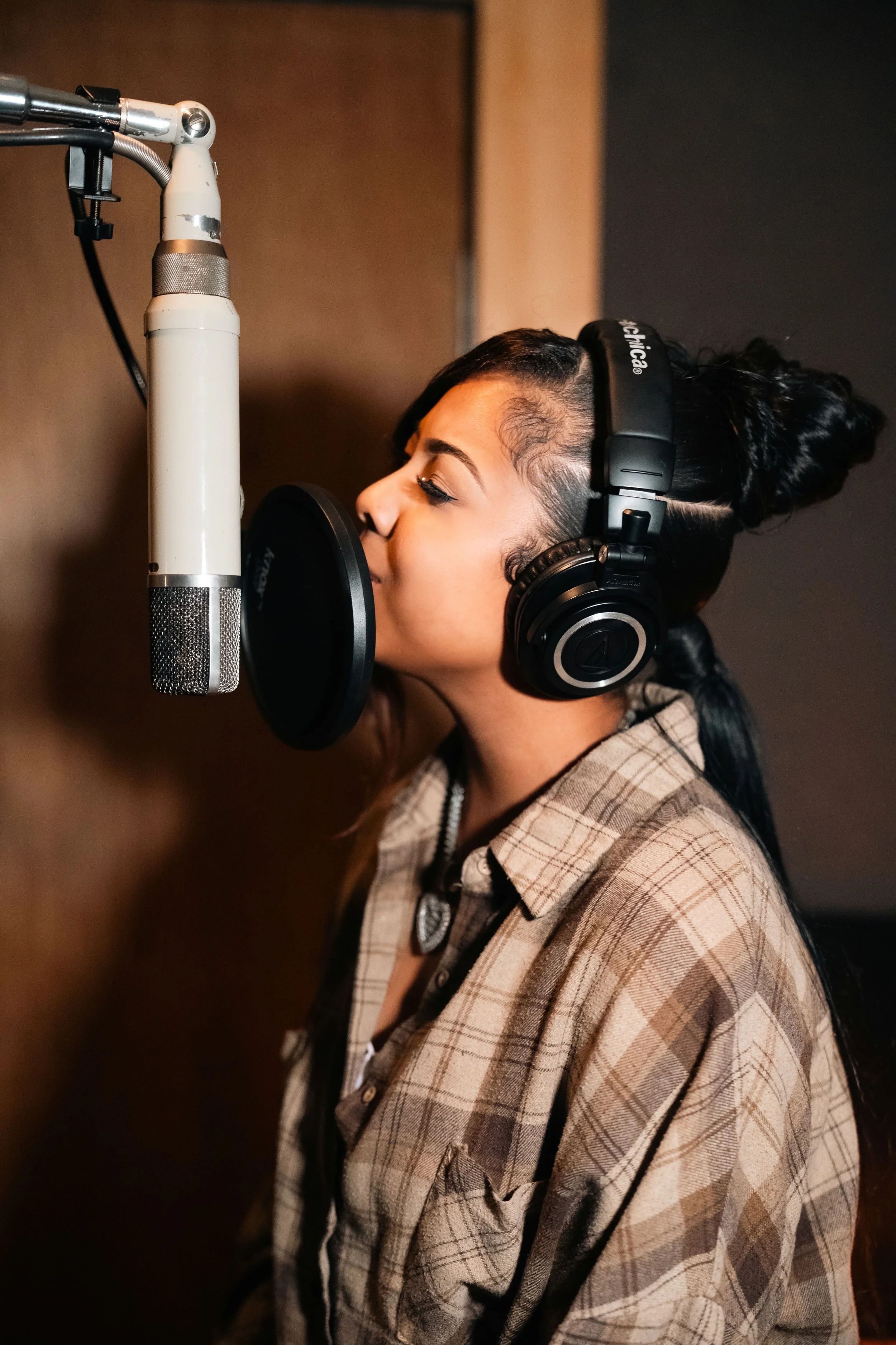 A woman in a plaid shirt is singing into a microphone in a recording studio, wearing large black headphones.