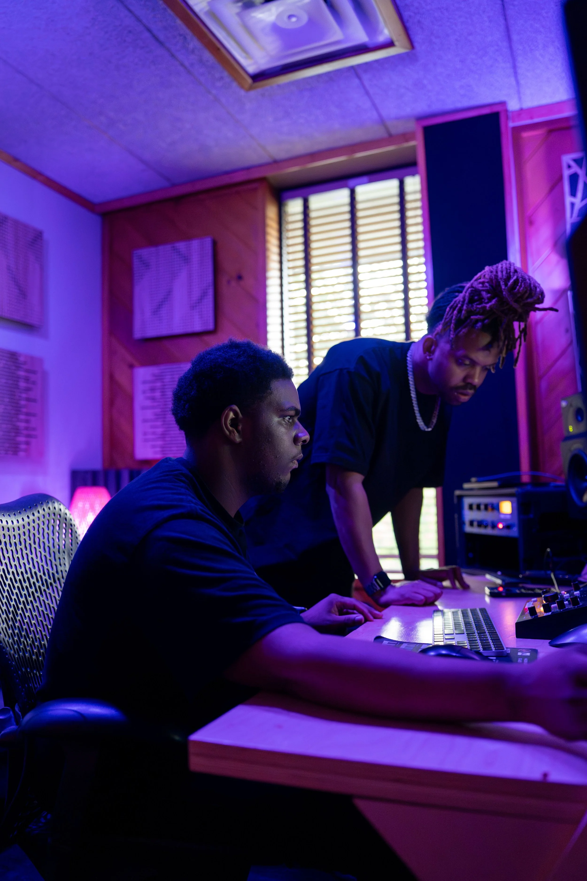 Two men in a music studio with purple and blue lighting, working with electronic music equipment, one seated at a desk and the other standing, with soundproof walls and window blinds in the background.