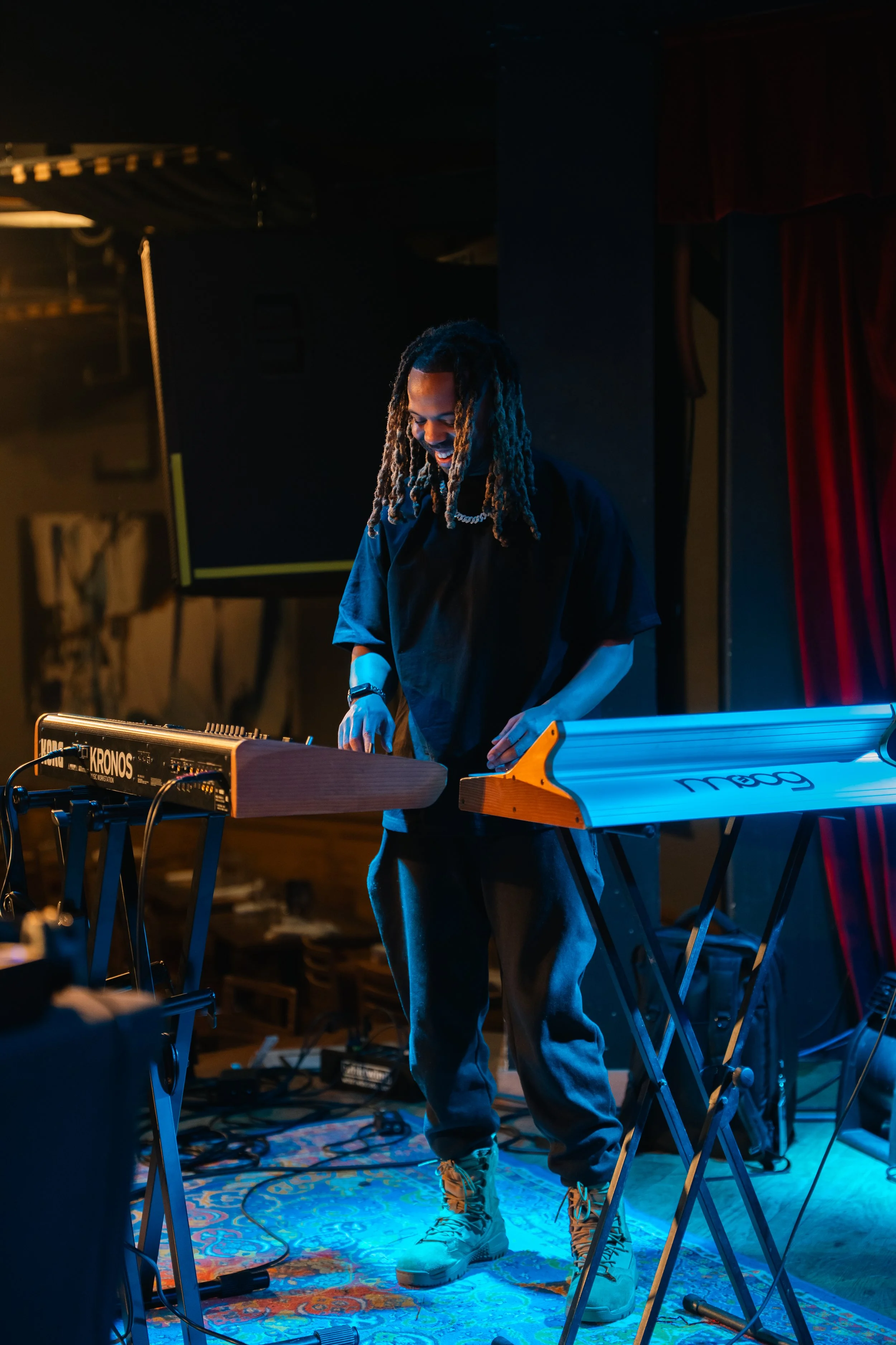 A musician with dreadlocks wearing a black shirt and gray pants standing behind a keyboard on a stage with colorful lighting and red curtains.